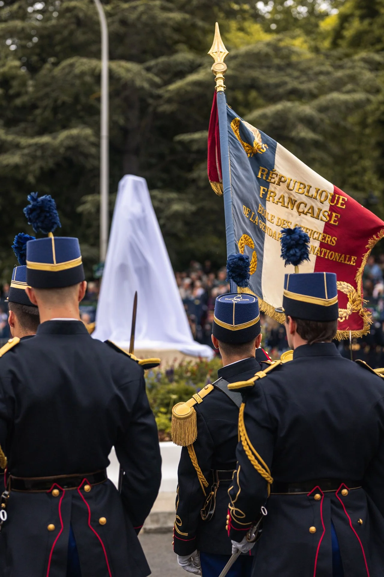 Soldats en uniforme noir avec des garnitures dorées participent à une cérémonie, avec un drapeau français. Le drapeau montre la République Française et un autre symbole. Il y a un cercueil drapé en blanc dans le fond, entouré de spectateurs.