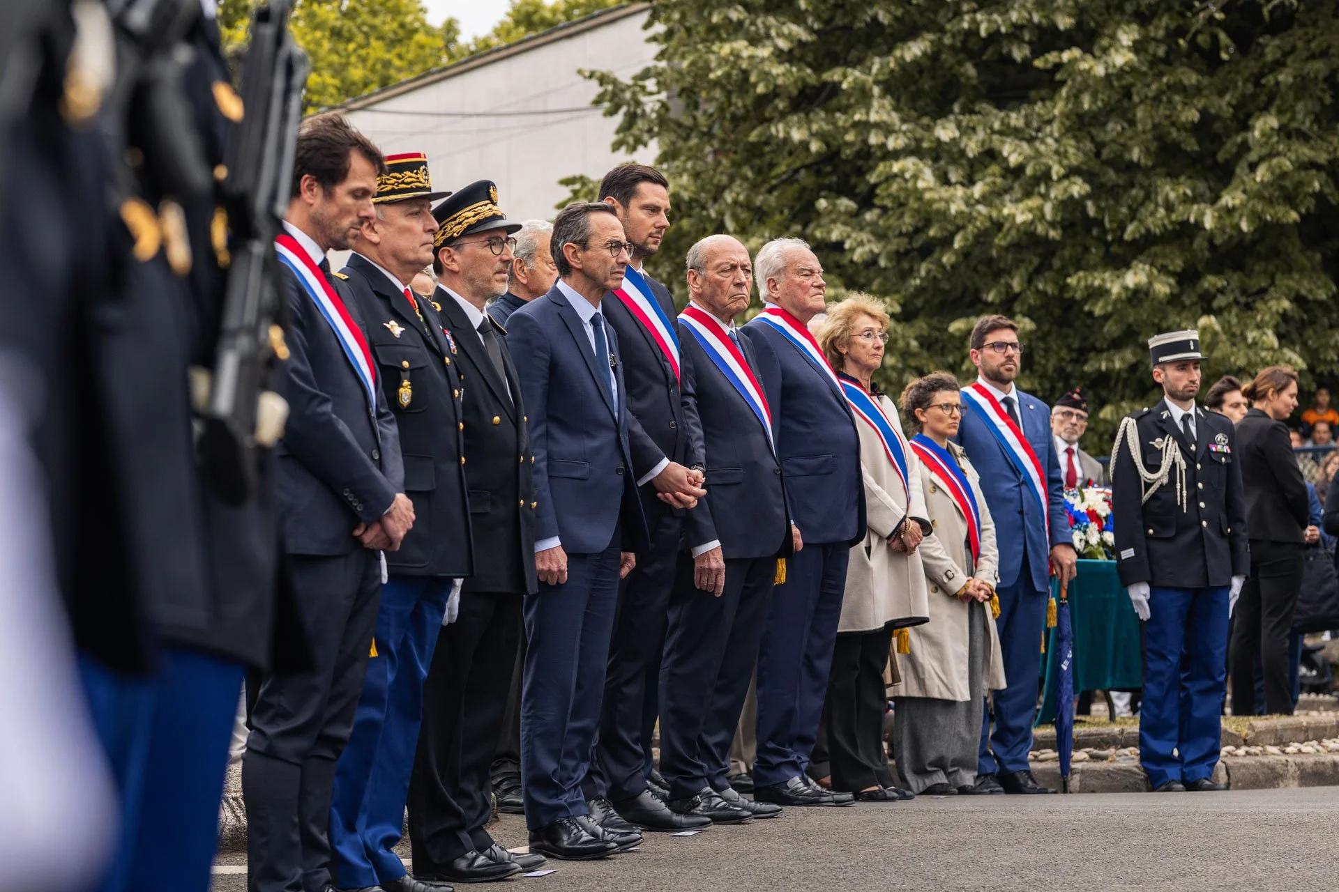Groupe de personnes en tenue officielle lors d'une cérémonie commémorative, portant laoon, en présence de militaires et de civils, sous un arbre vert avec un bâtiment en arrière-plan.