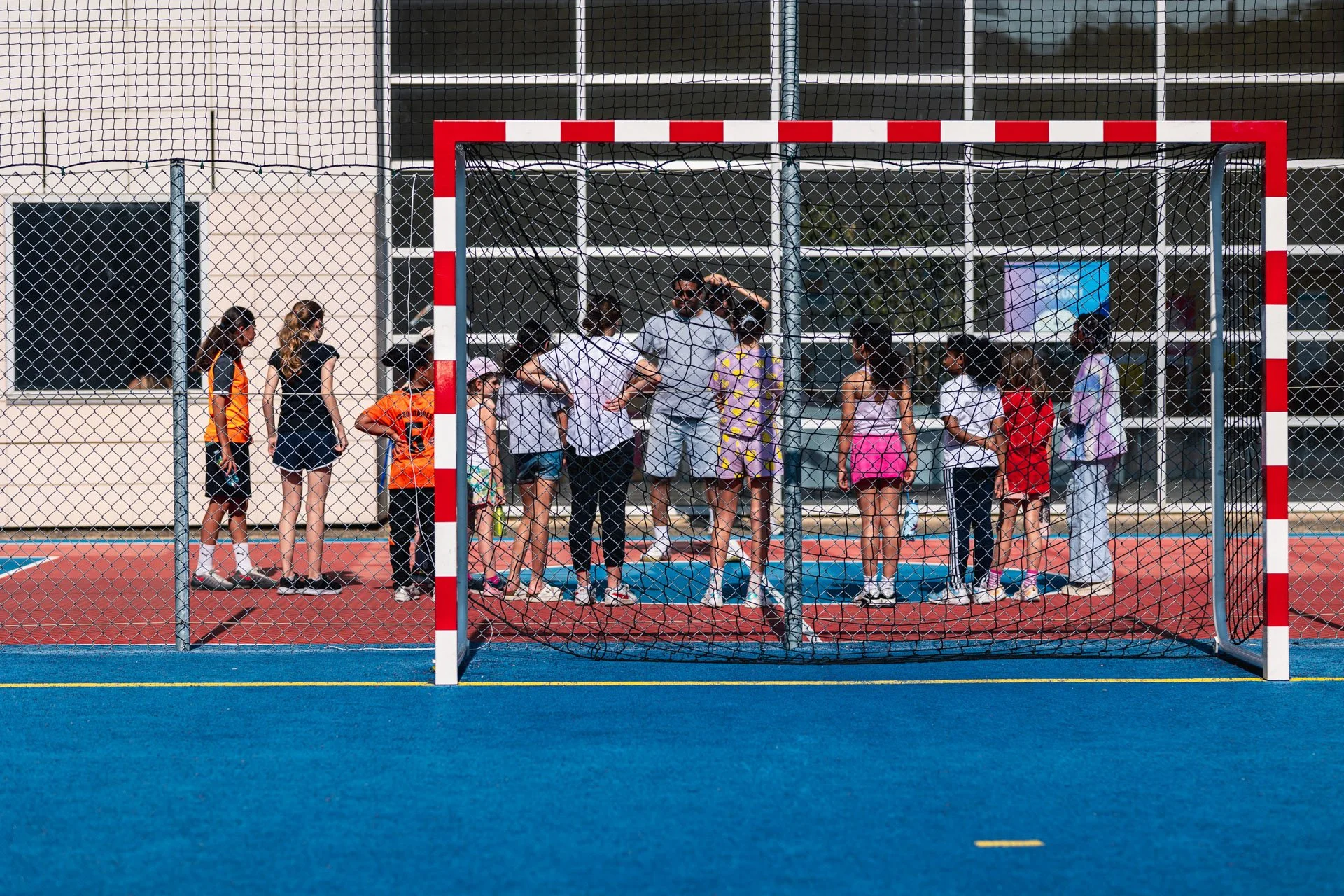 Un groupe d'enfants et d'adultes jeunes, derrière une clôture de terrain de sport, en train de participer à une activité ou un événement en plein air, avec un bâtiment en arrière-plan.