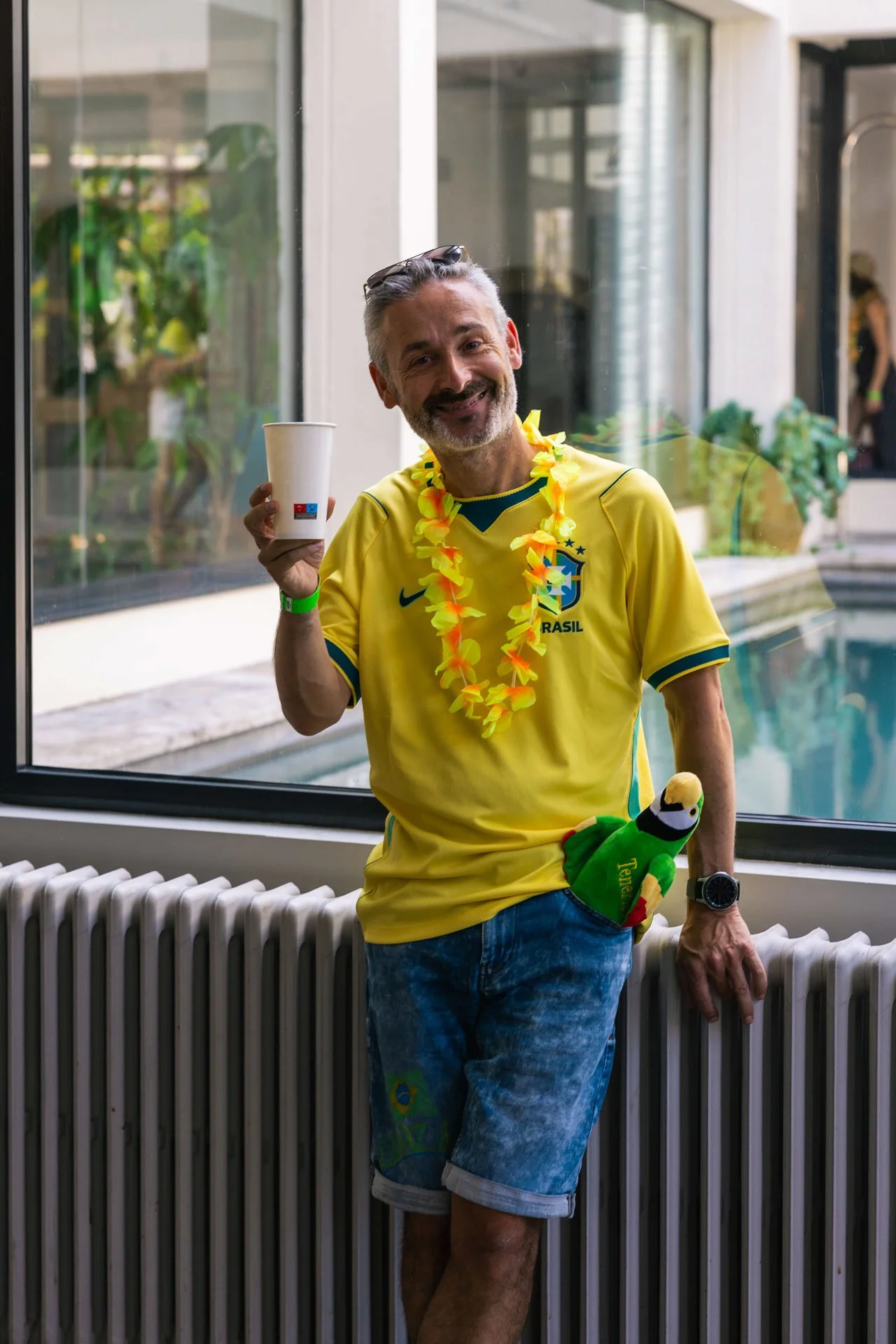 Un homme souriant porte un maillot jaune du Brésil, une lei hawaïenne, une montre, et tient une poupée de football vert et jaune derrière une fenêtre.