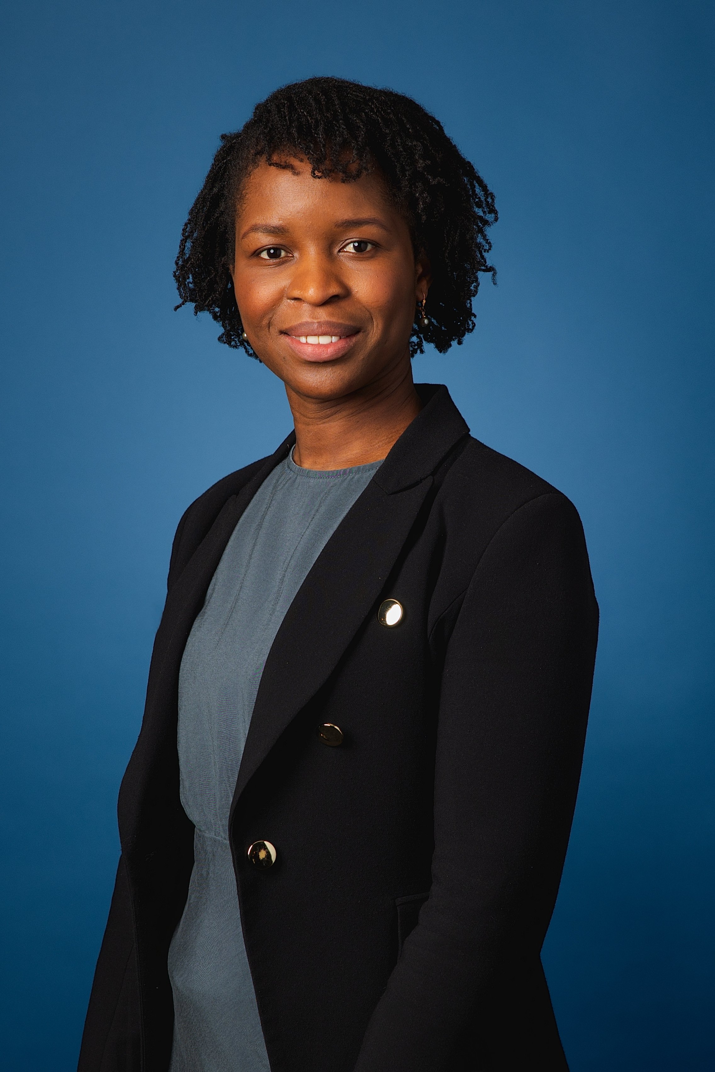 Portrait d'une femme noire portant un blazer noir et une robe grise, avec un fond bleu.