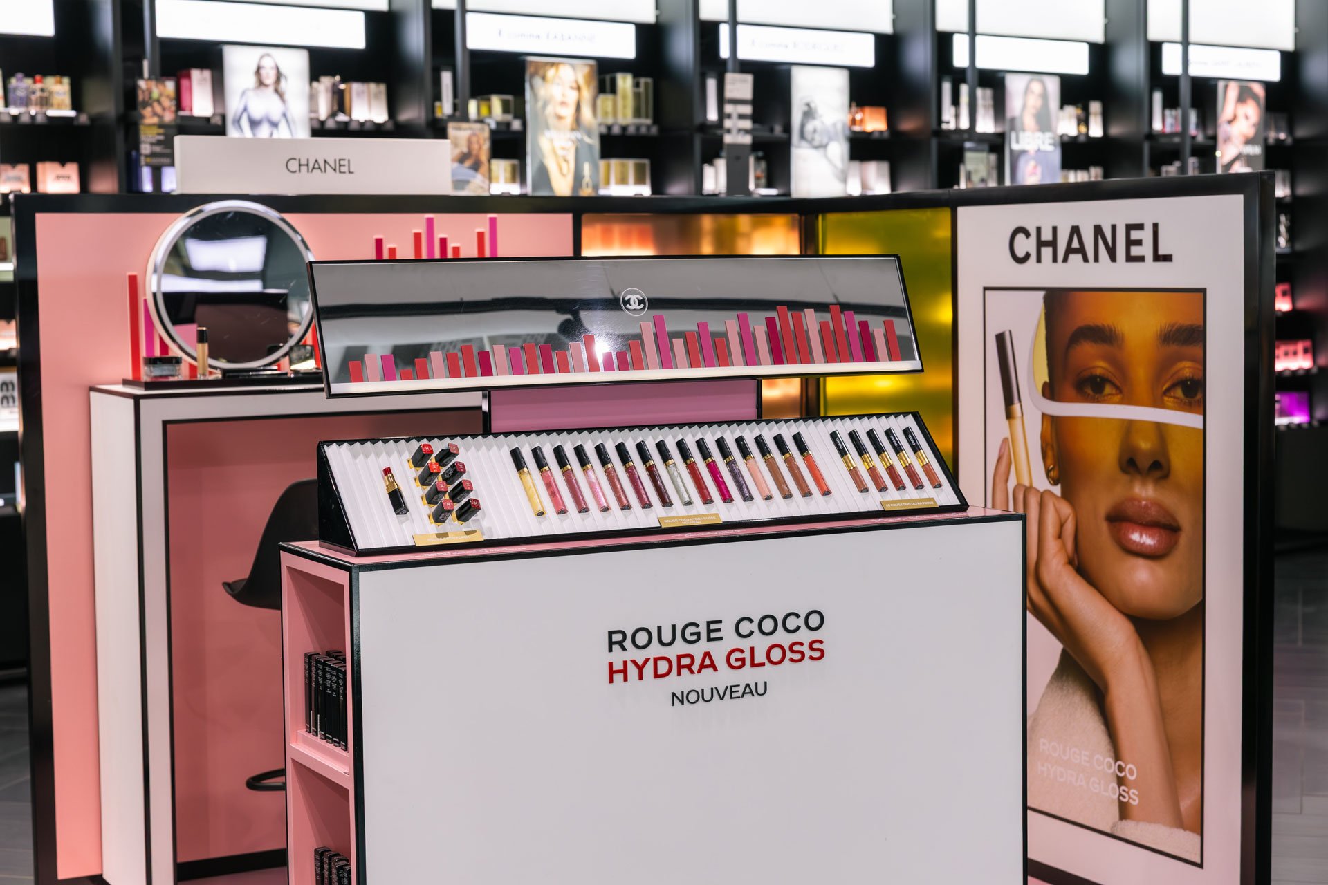 Présentoir de la marque Chanel avec des produits de maquillage, notamment des rouges à lèvres, dans un magasin. Une affiche promotionnelle avec une femme portant un maquillage sophistiqué est visible en arrière-plan.