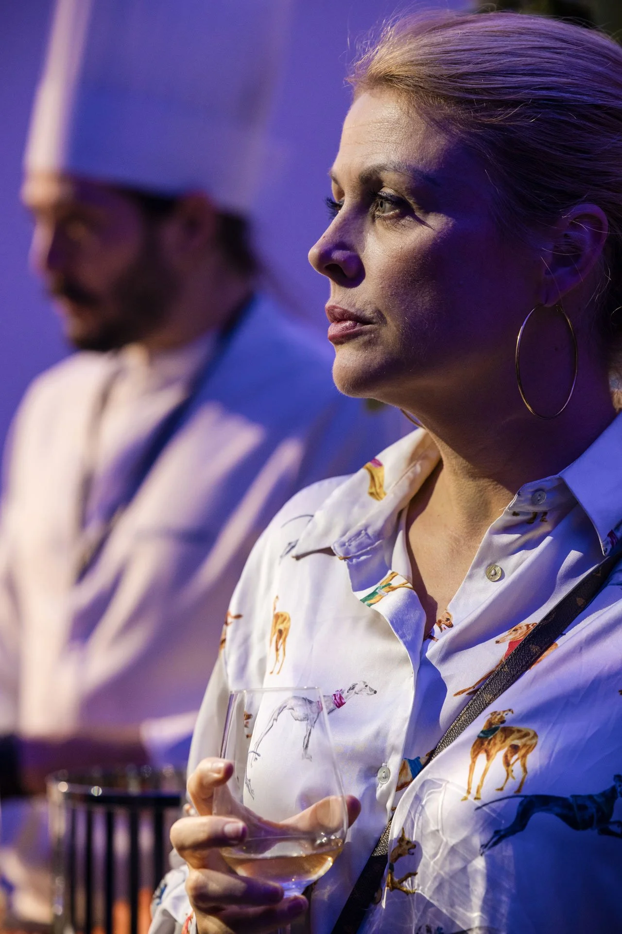Une femme avec des cheveux blonds portant une chemise blanche à motifs de girafe, tenant un verre de vin blanc, lors d'un événement ou d'une dégustation.