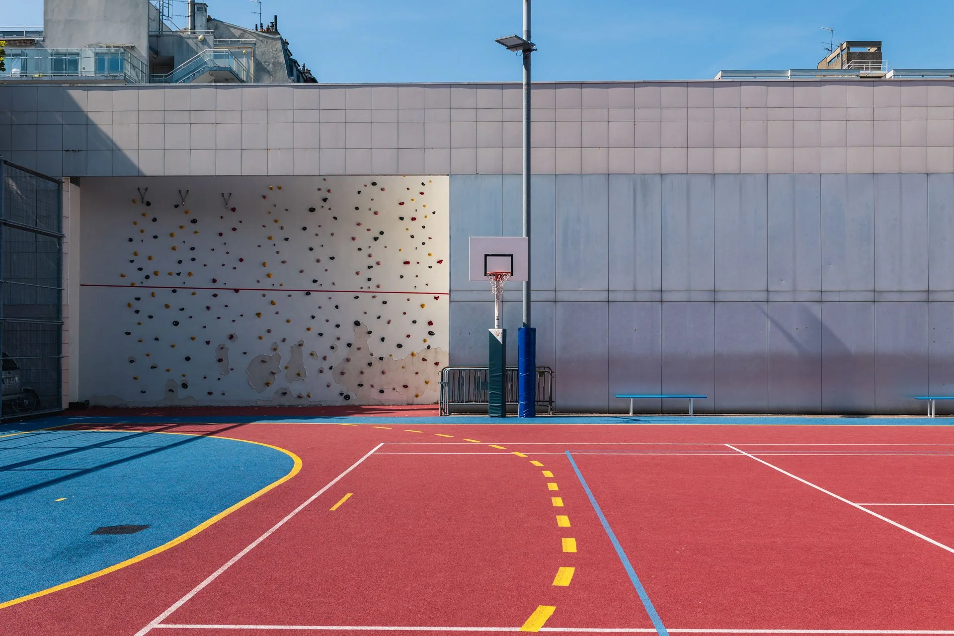 Un terrain de basketball extérieur avec un mur d'escalade à gauche et un panneau d'affichage avec un panier de basketball au centre. Le sol est coloré en rouge, bleu et jaune, avec des lignes blanches et jaunes délimitant les zones de jeu.