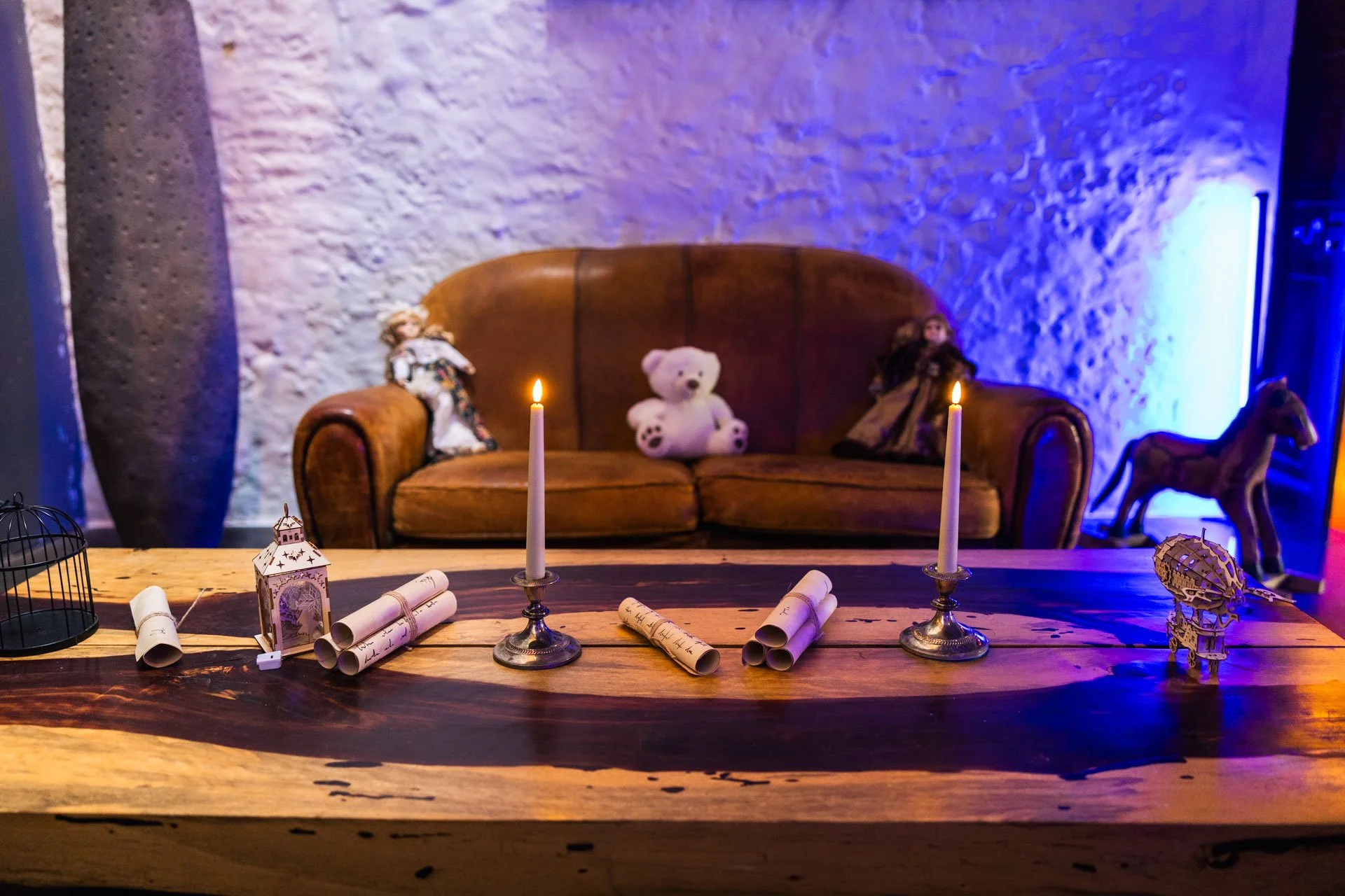 Table en bois avec des bougies, des rouleaux de papier, une cage d'oiseau, une lanternes, des objets décoratifs et un canapé en cuir en arrière-plan avec des poupées en peluche et des jouets en bois, éclairé par des lumières douces et colorées.