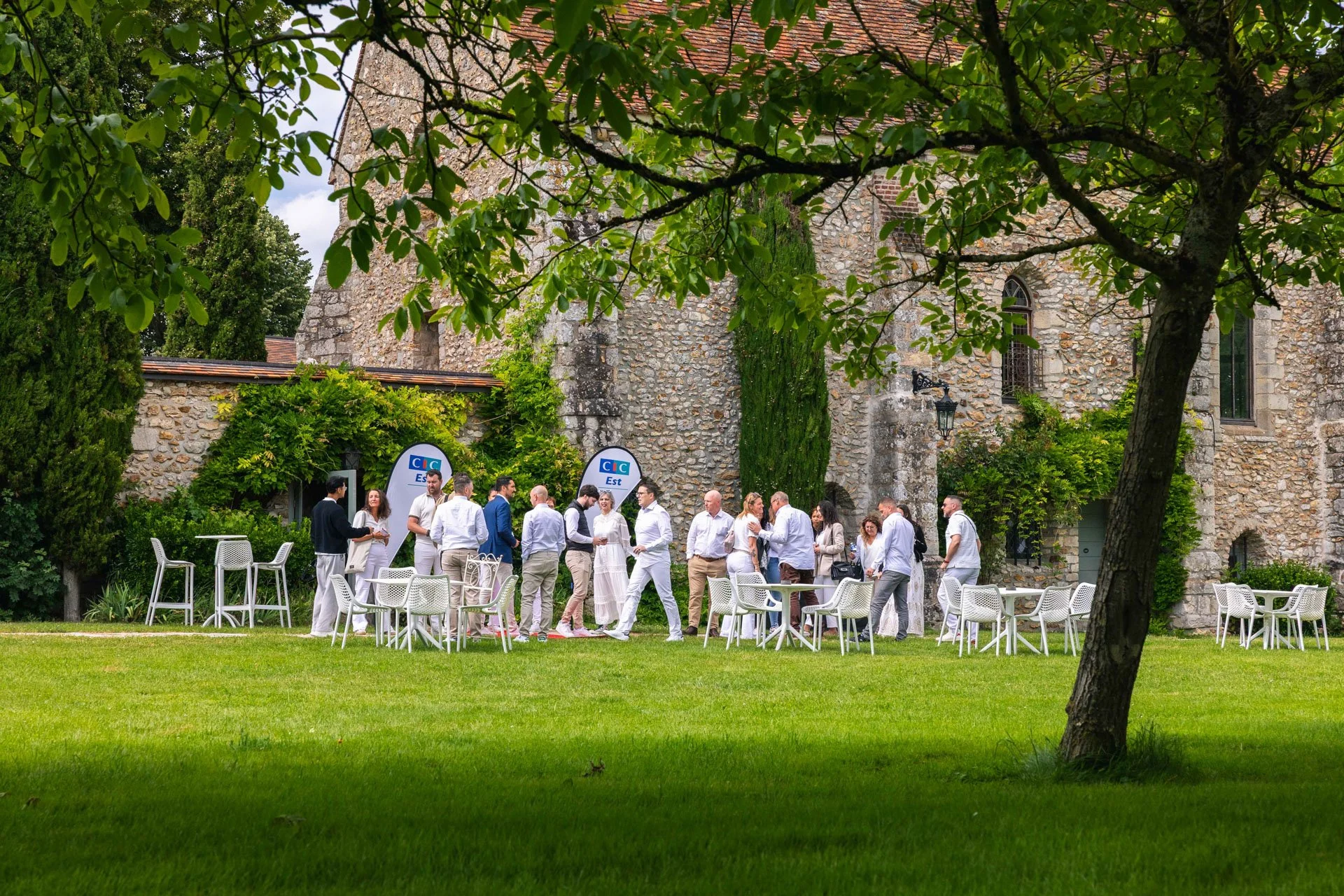 Groupe de personnes en tenue décontractée socialisant dans un jardin, avec une vieille bâtisse en pierre en arrière-plan, entourée d'arbres et de mobilier de jardin blanc.
