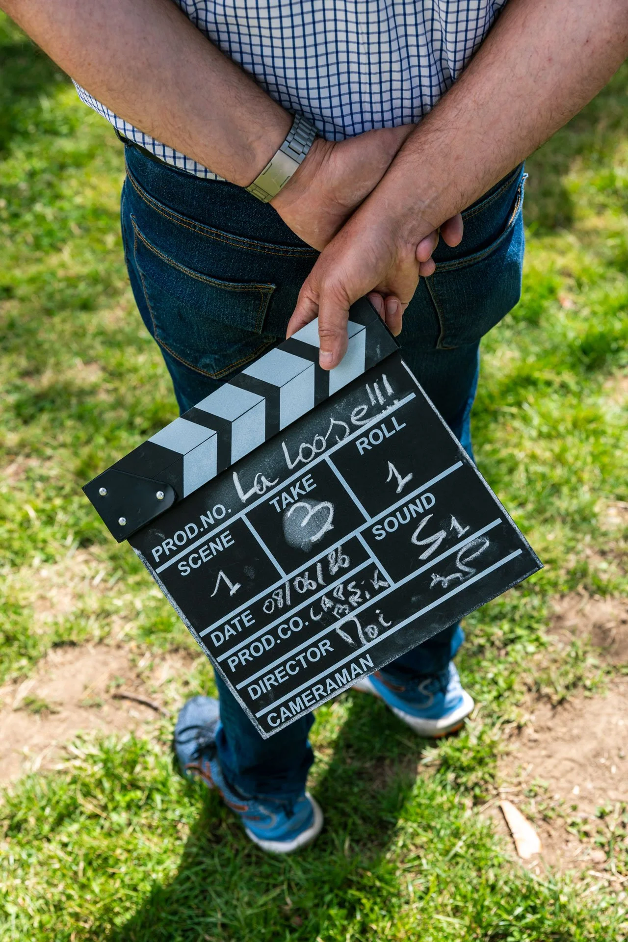 Personne tenant une planche de clap de cinéma avec un foulard autour du cou et des chaussures de sport, marché sur de l'herbe lors d'un tournage.