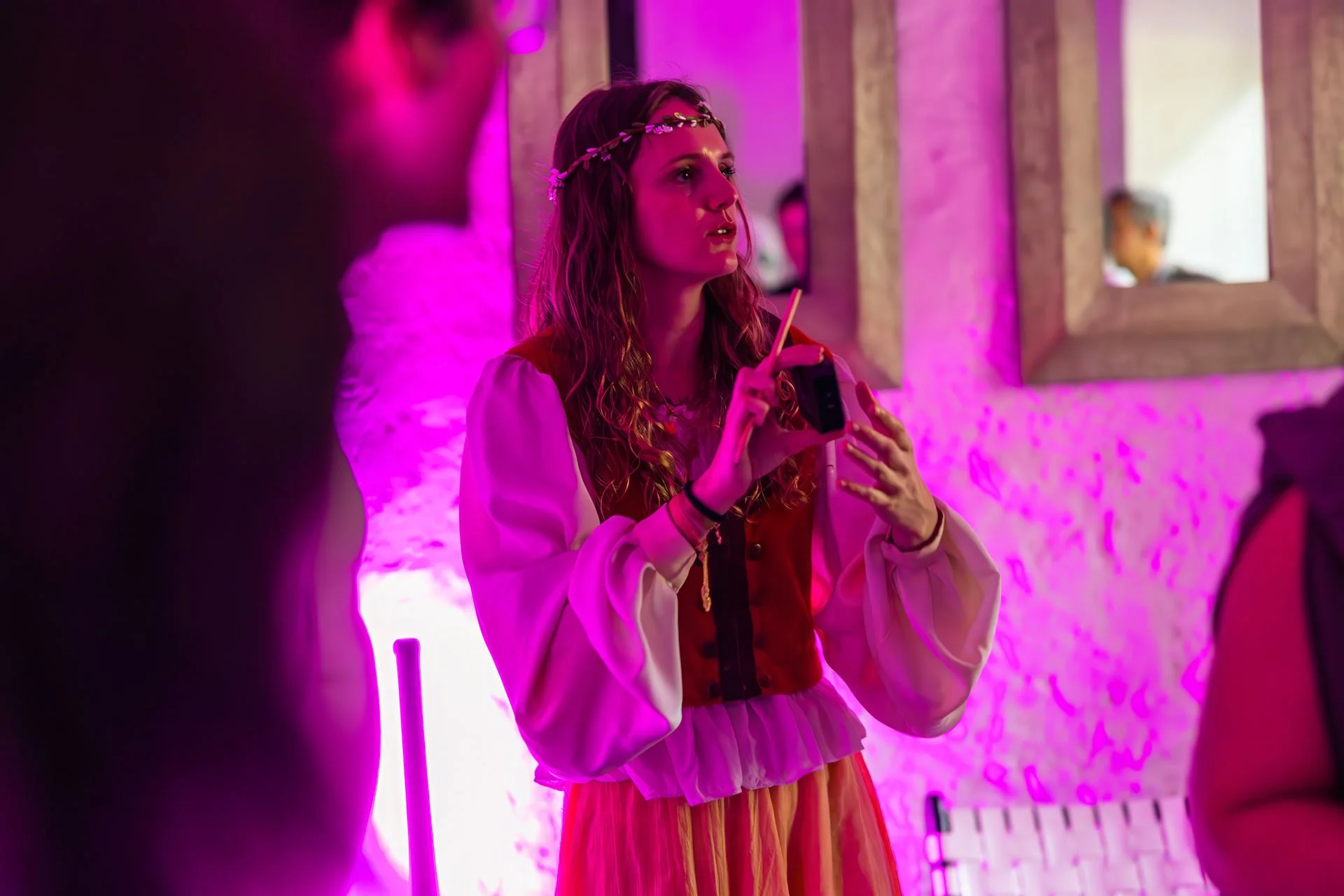 Une jeune femme avec une couronne de fleurs discute dans une pièce éclairée en rose, portant une veste rouge et une jupe longue à rayures. Elle tient un téléphone portable.