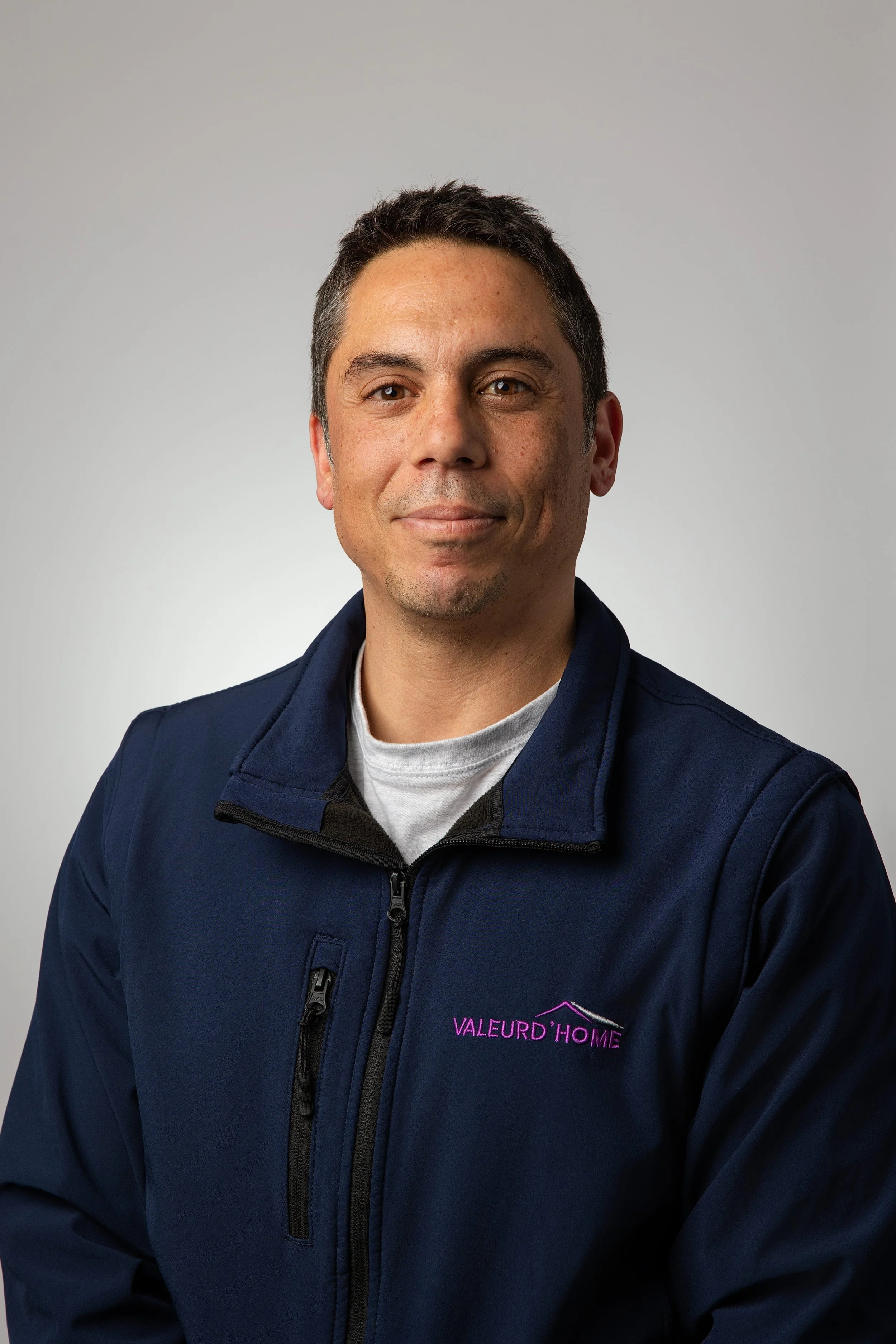 Un homme porte une veste noire avec un logo violet 'VALEURD HOME' et regarde l'appareil photo, souriant légèrement, seul dans le cadre.