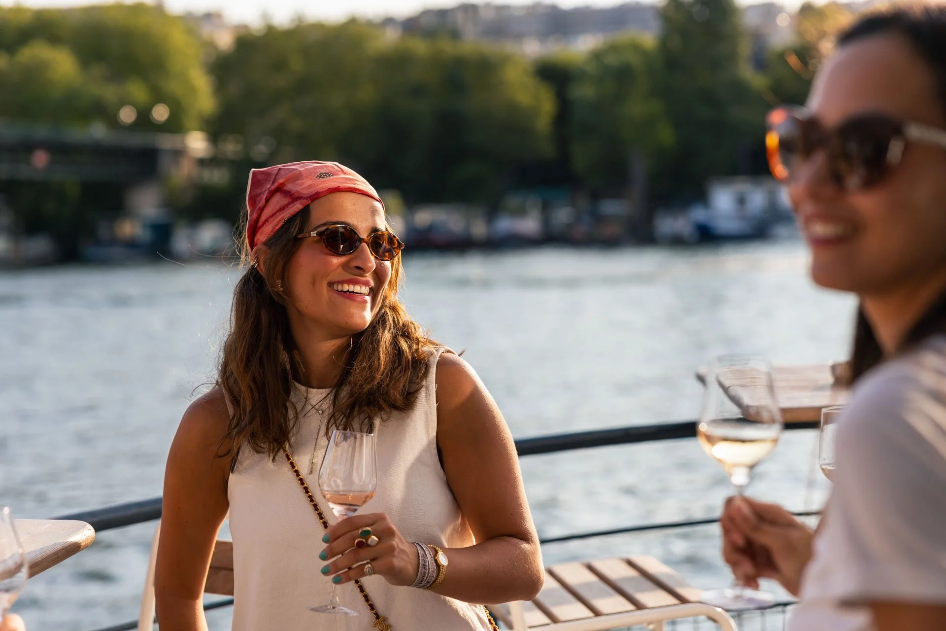 Deux femmes souriantes portant des lunettes de soleil, chacune tenant un verre de vin rosé lors d'une sortie en bateau, avec un paysage fluvial et des arbres en arrière-plan.