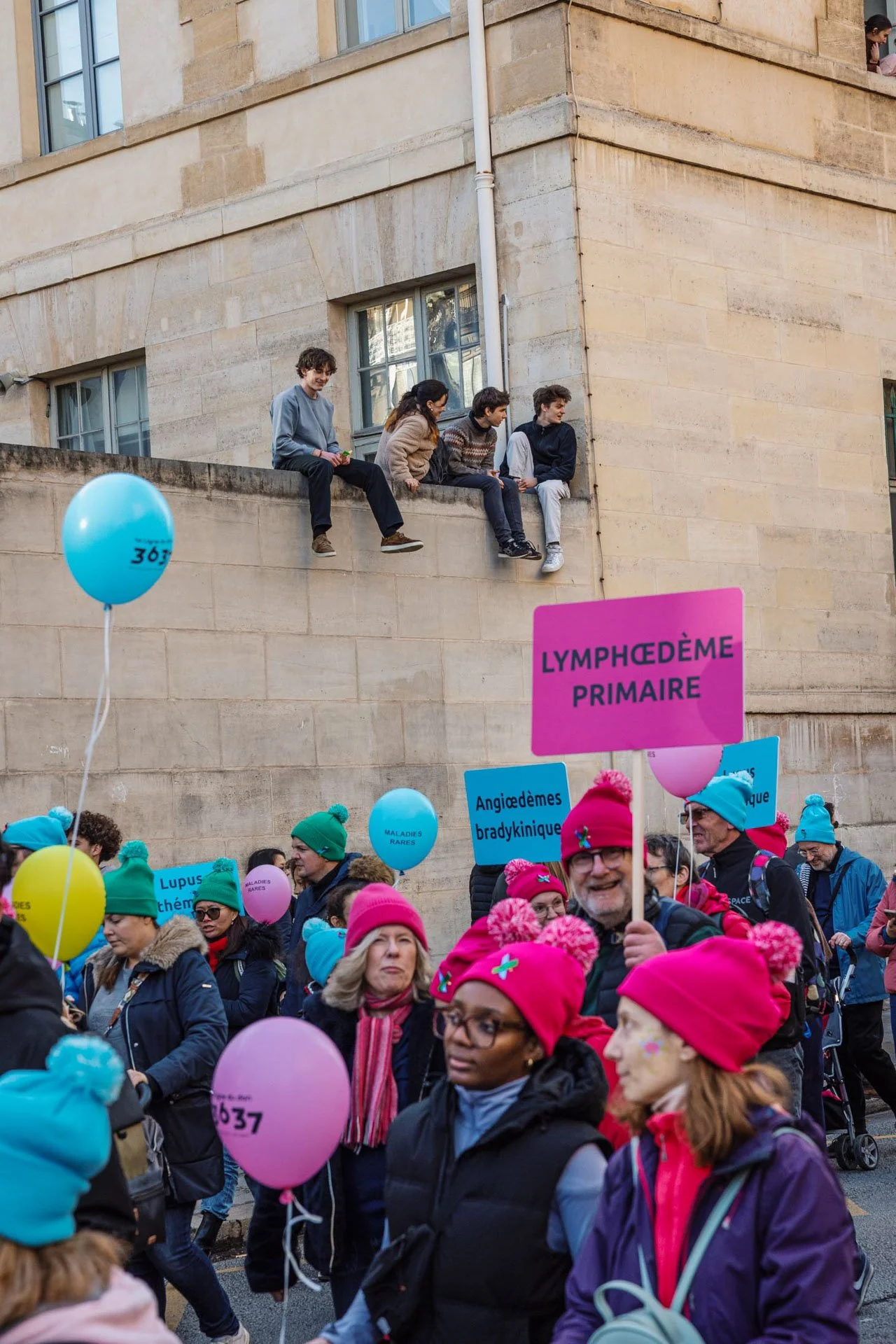 Manifestation pour sensibiliser à la lymphœdème primaire avec des panneaux et des ballons colorés, des personnes portant des chapeaux rose et des vêtements chauds dans la foule.