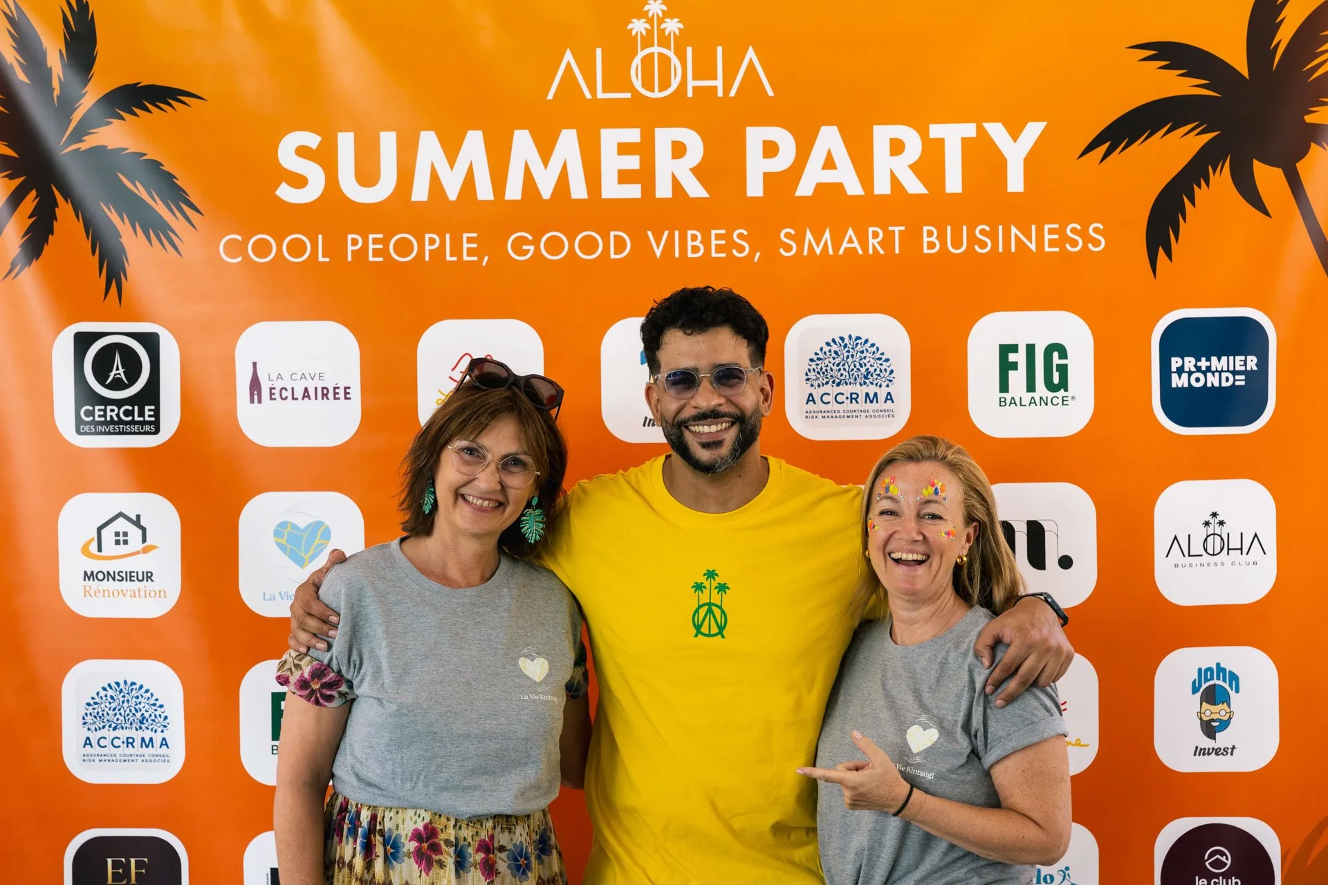 Photo de trois personnes souriantes posant devant un fond orange avec des logos d'entreprises et de sponsors lors d'un événement intitulé 'Summer Party' avec le slogan 'Cool people, good vibes, smart business'.