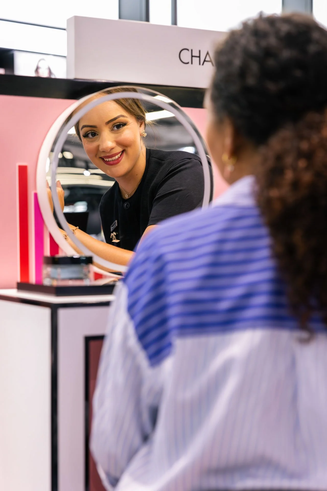 Une femme regarde son reflet dans un miroir à une vitrine de parfumerie, avec un panneau portant le nom "Chanel" en arrière-plan.