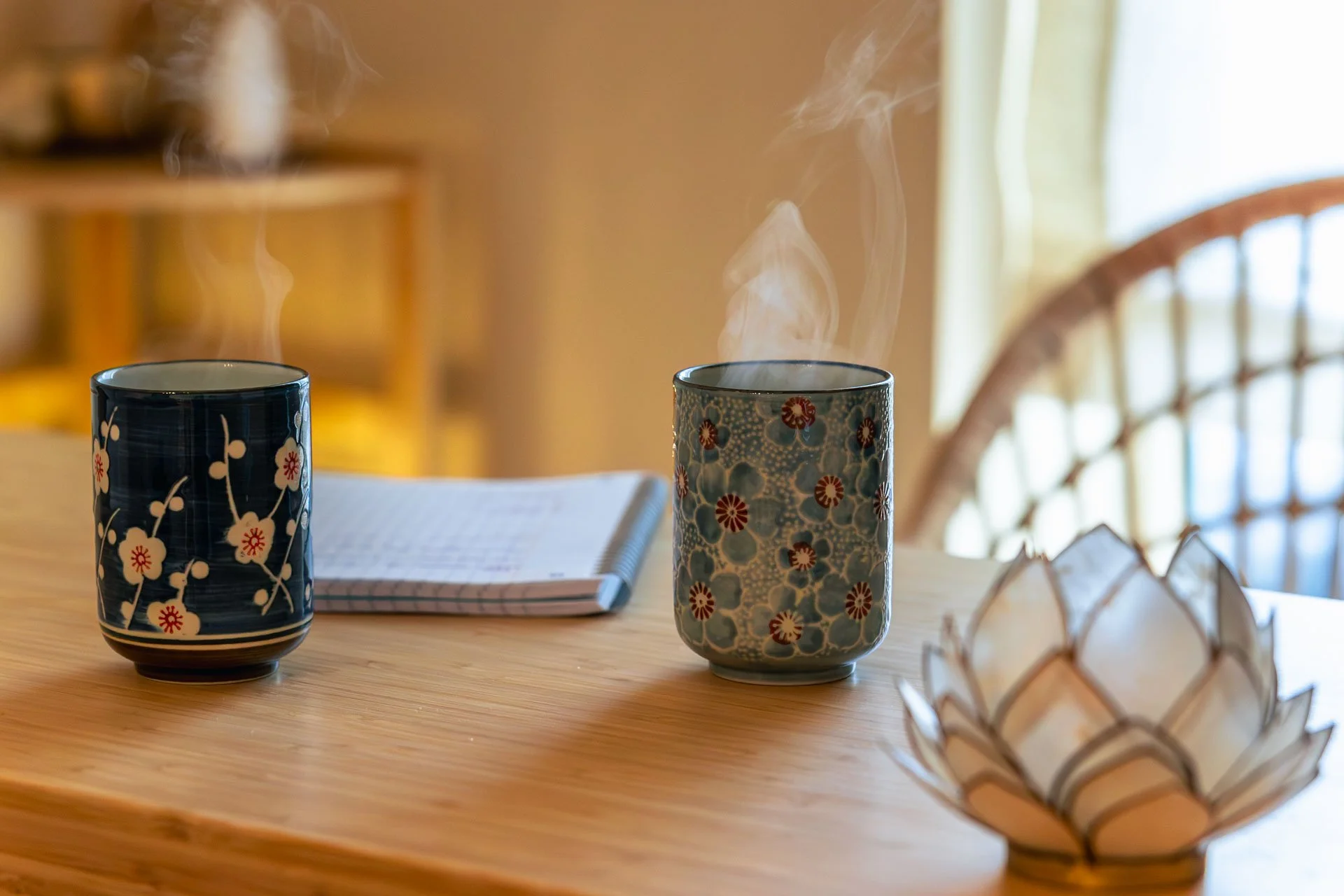 Deux tasses à café en céramique avec des motifs floraux, posées sur une table en bois, avec un carnet ouvert derrière elles. Dans le coin droit, un objet décoratif en forme de panier en fil de fer est visible.
