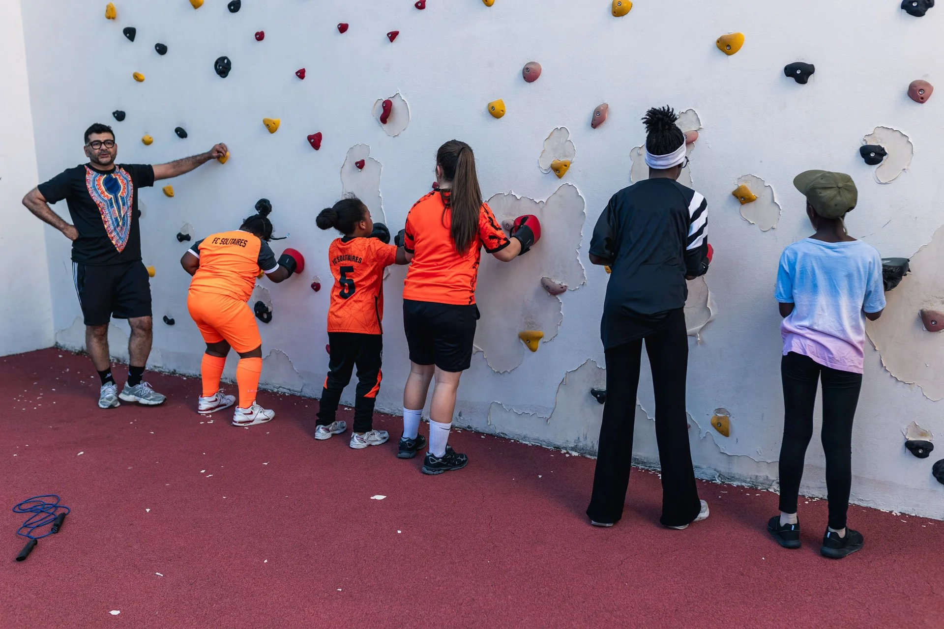 Un groupe de jeunes fait de l'escalade sur un mur d'escalade en extérieur, sous la supervision d'un adulte.