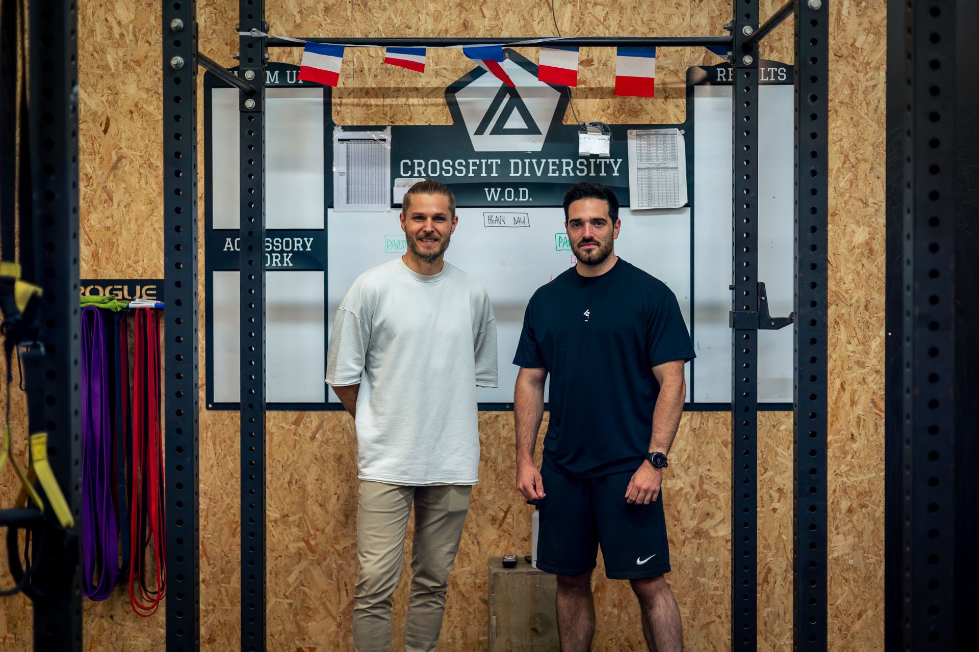 Deux hommes posant dans une salle de sport CrossFit, avec un mur en contreplaqué derrière eux et des équipements d'entraînement à gauche, dont des bandes de résistance colorées. Un panneau indique 'CrossFit Diversity'.