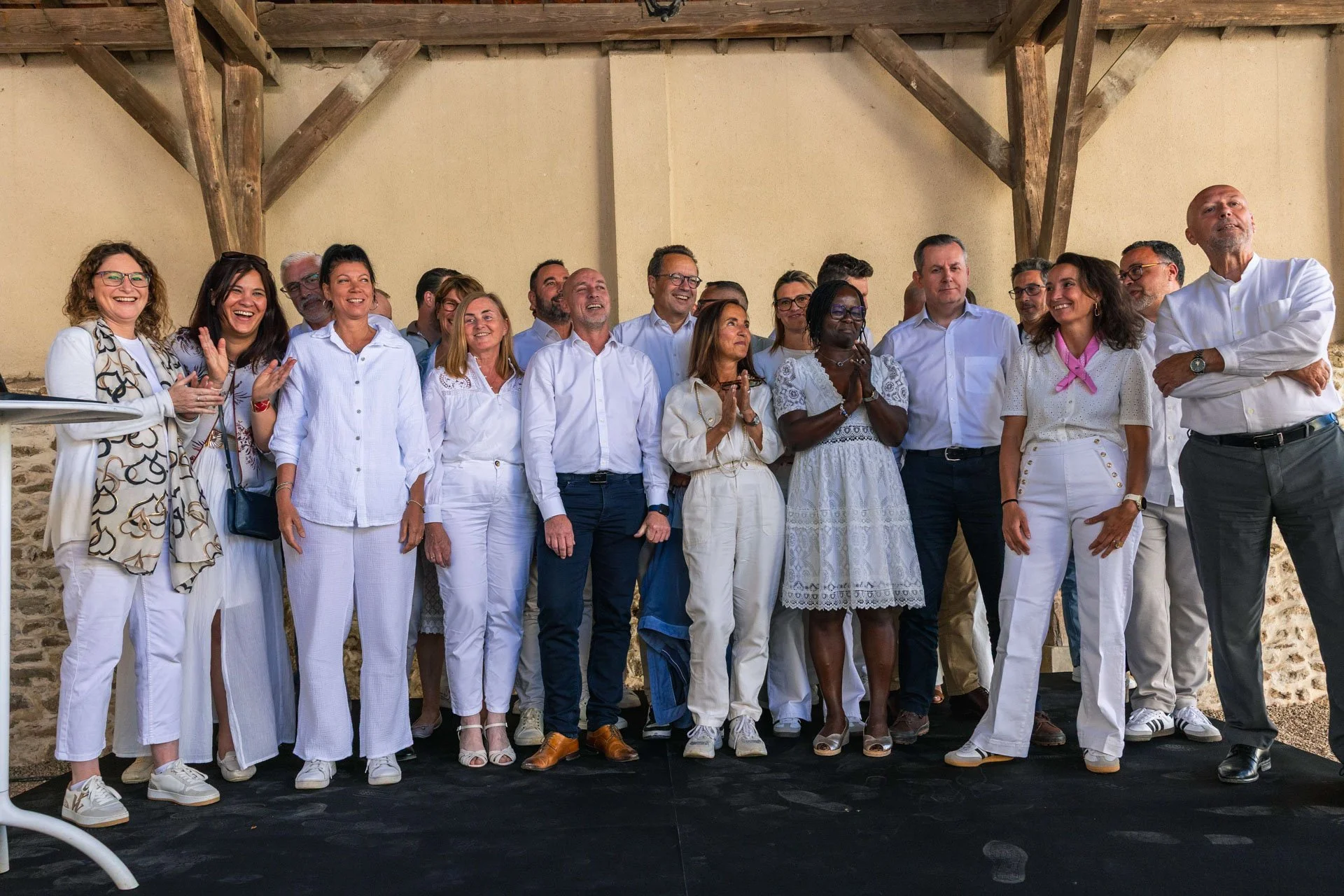 Groupe de personnes debout, souriantes, portant des vêtements blancs, lors d'un événement en intérieur, sous une structure en bois.