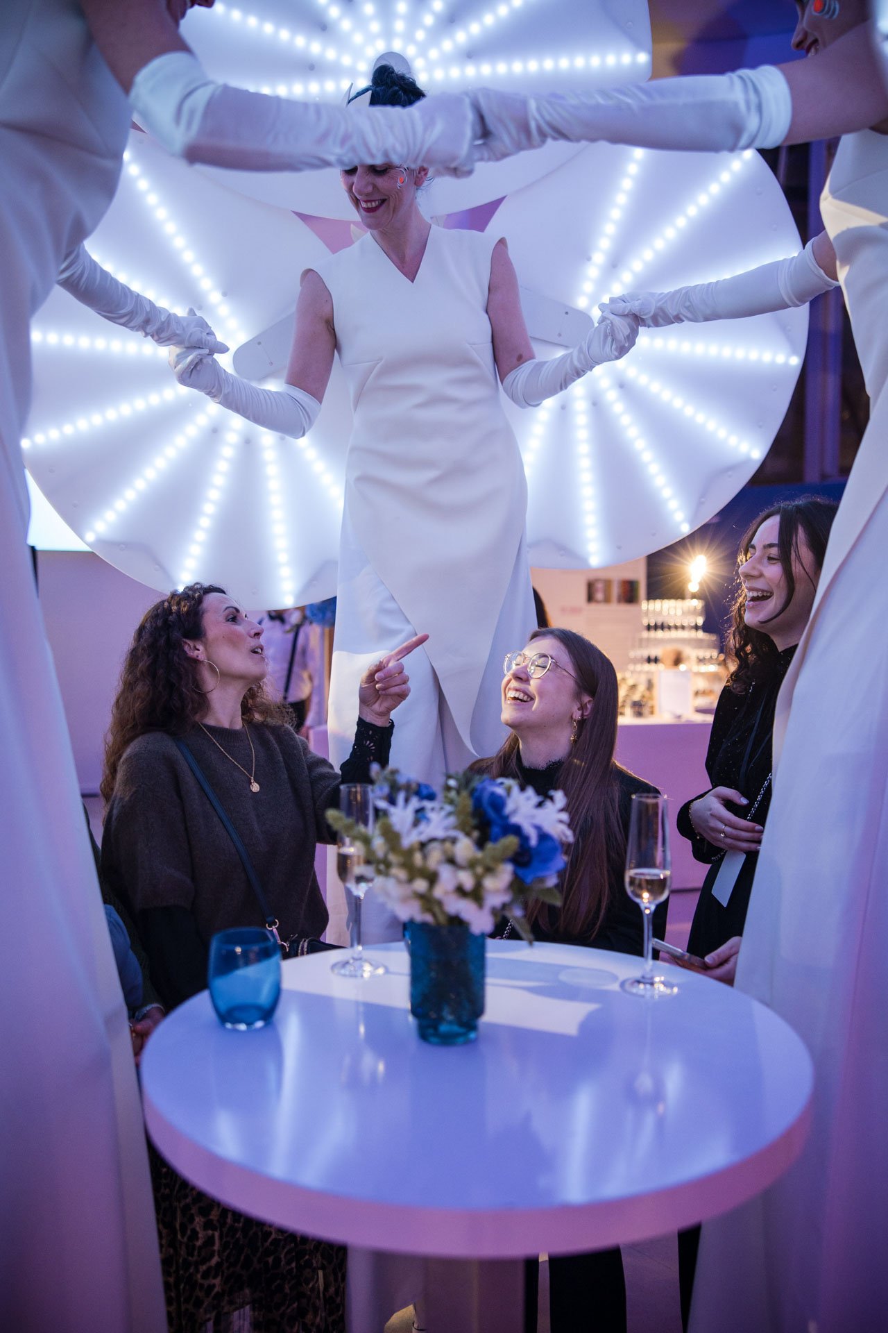Groupe de femmes assises autour d'une table lors d'une fête ou d'un événement, avec une femme debout au centre, décor éclairé avec des lampes LED, ambiance joyeuse.