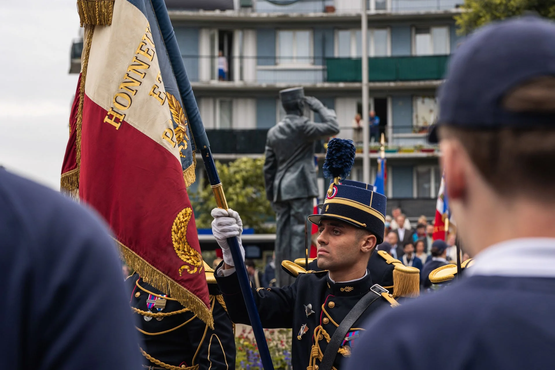 Soldat français en uniforme noir et or tenant un drapeau français lors d'une cérémonie, avec une statue de soldat en arrière-plan et des spectateurs en arrière-plan.