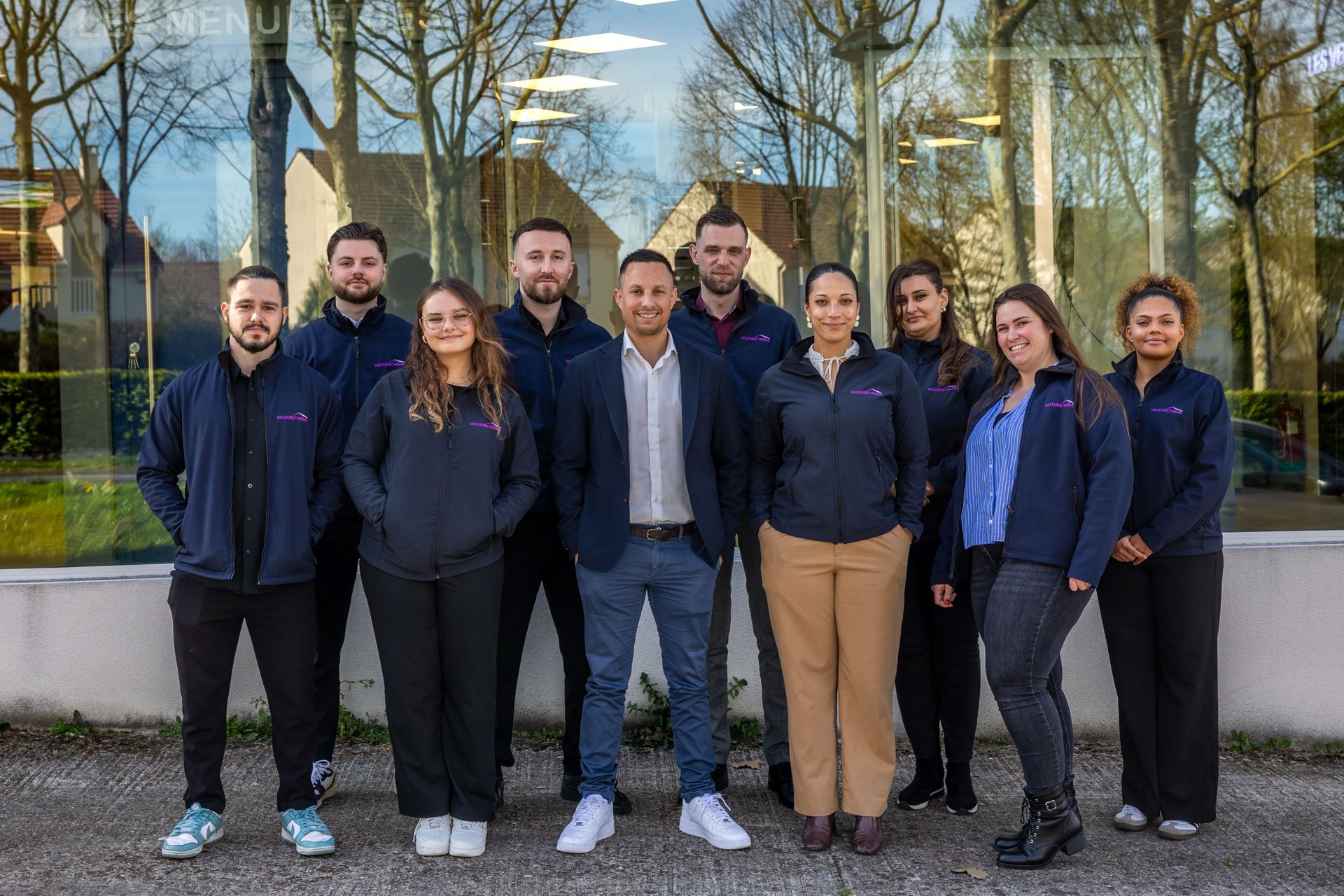 Groupe de dix personnes posant devant une vitrine avec réflexion d'arbres et de maisons, certaines en vêtements de travail avec logo violet, d'autres en tenues professionnelles, souriantes.
