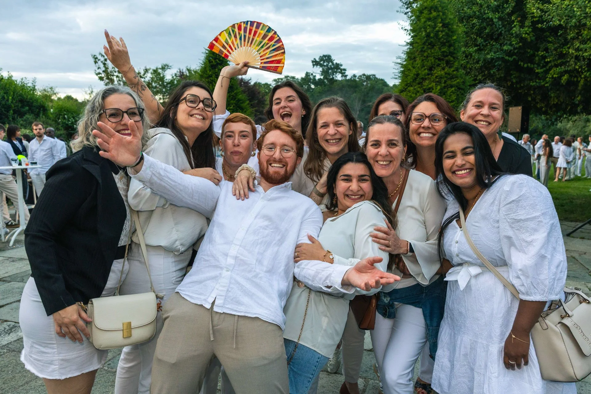 Groupe de personnes souriantes lors d'une fête en plein air, portant des vêtements blancs et certains avec des lunettes, avec des arbres en arrière-plan.