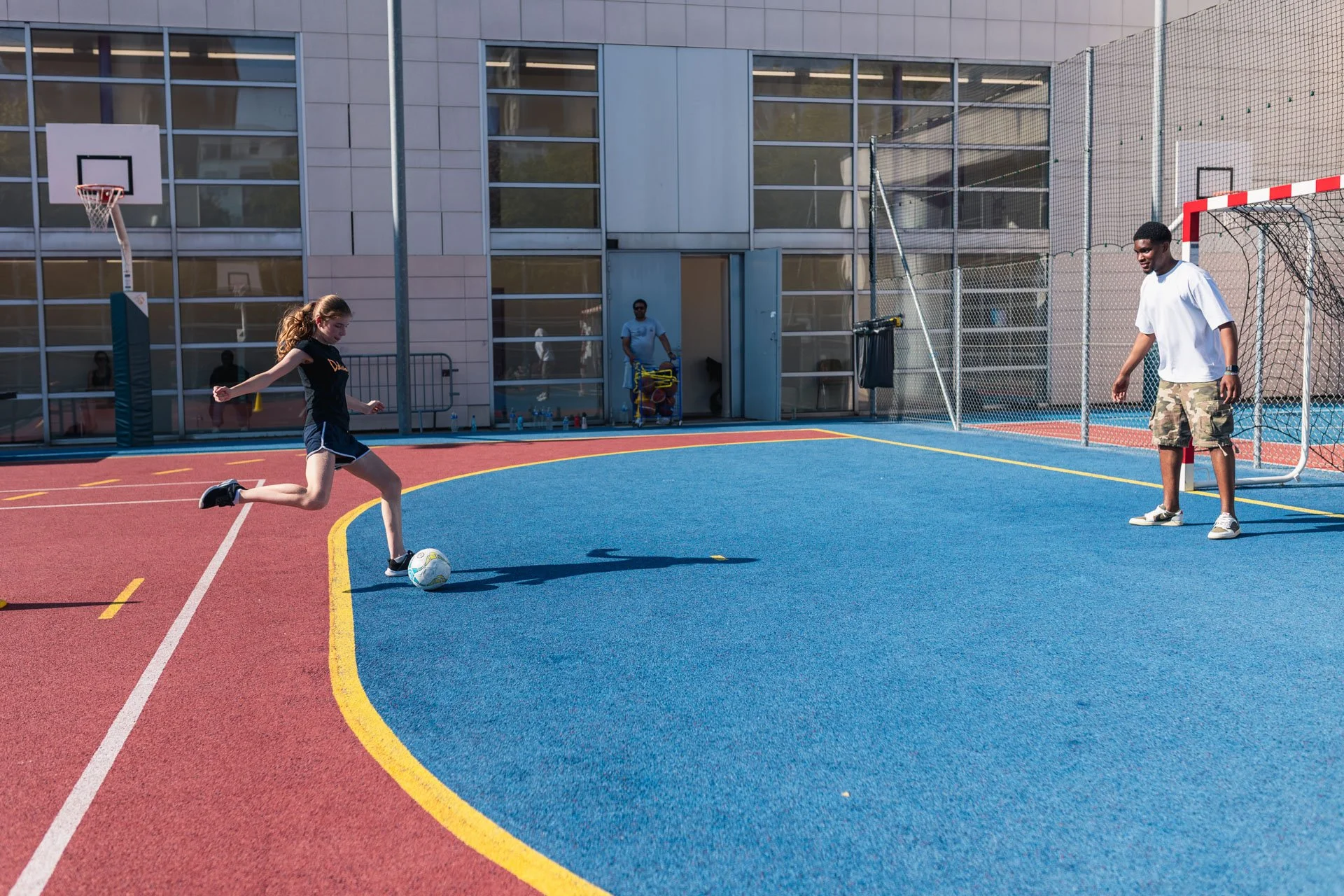 Une jeune fille joue au football sur un terrain en plein air, avec un gardien de but en face, deux but, et un bâtiment moderne en arrière-plan.