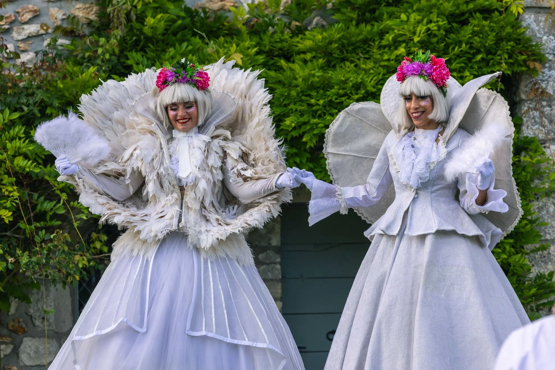 Deux femmes déguisées en costumes blancs élégants et ornés de fleurs roses et vertes, portant des coiffes de fleurs, dans un jardin avec des buissons verts et un mur en pierre en arrière-plan.