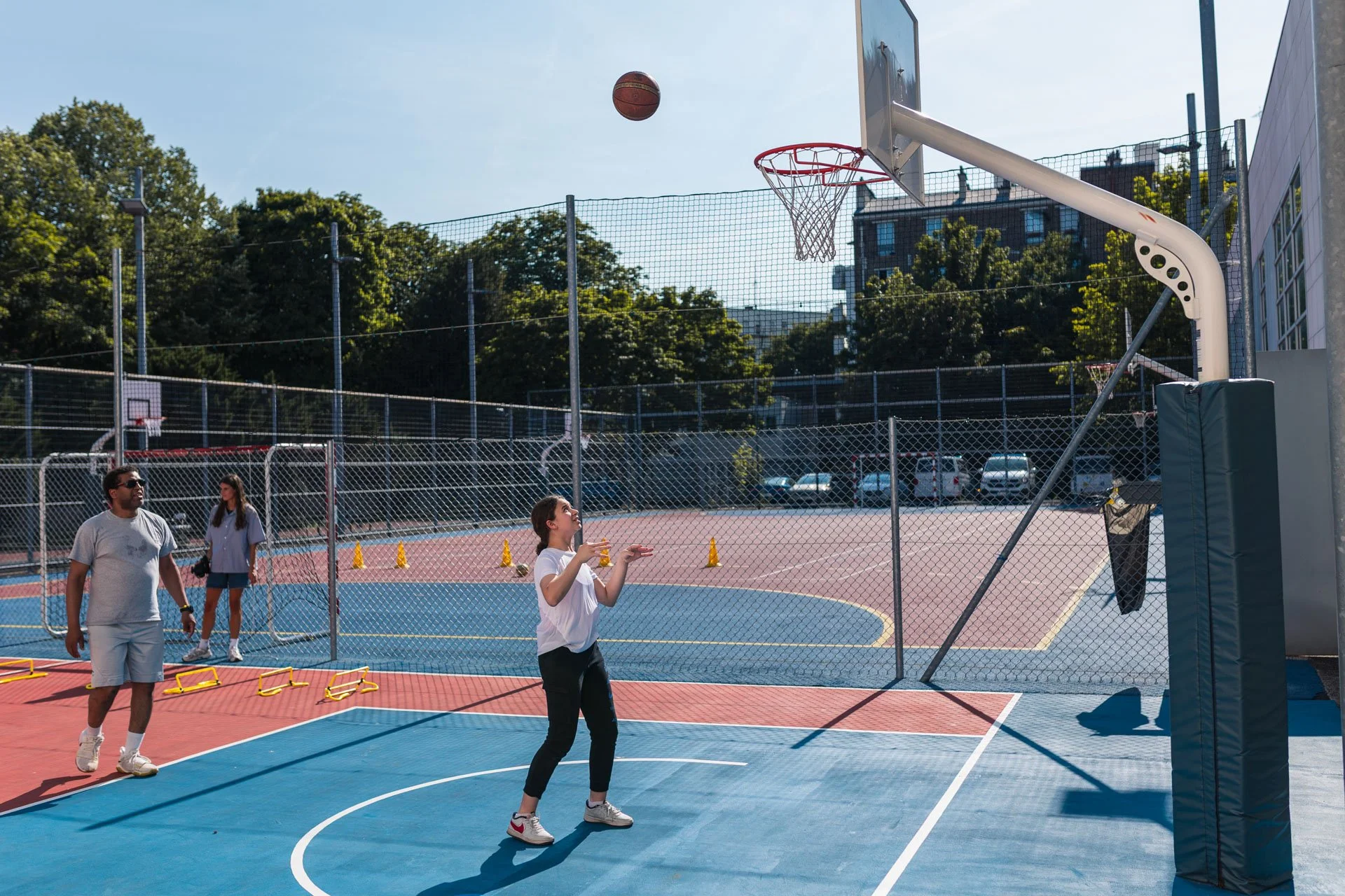 Jeune fille jouant au basketball sur un terrain extérieur sous un ciel ensoleillé, avec plusieurs personnes regardant ou attendant leur tour, entouré de filets et de barrières.