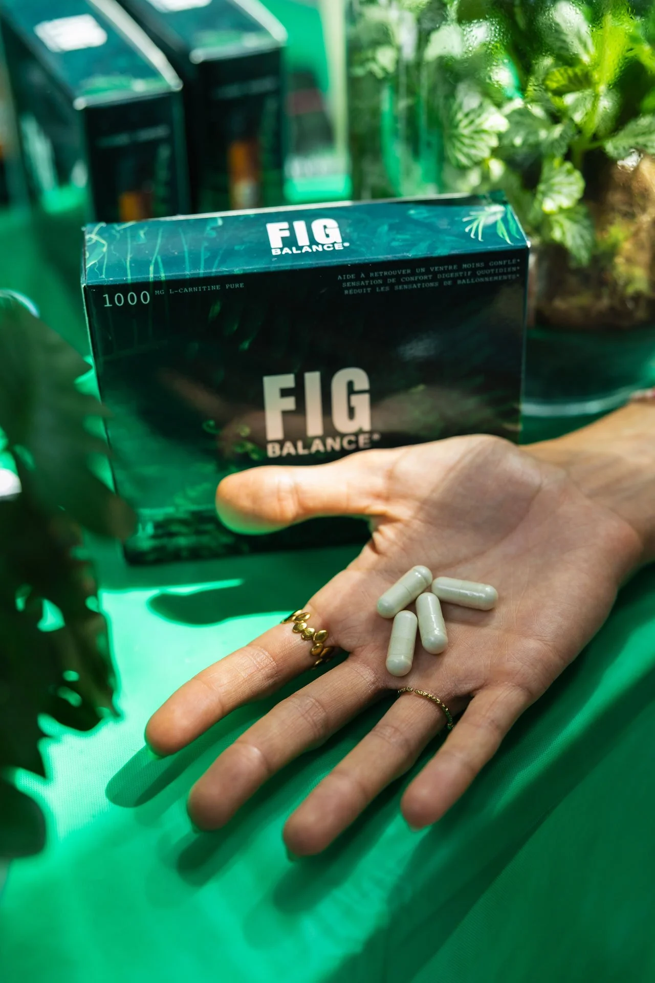 Main tenant des capsules de complément alimentaire blanches, avec un emballage de la marque 'FIG BALANCE' en arrière-plan avec des capsules sur un tapis vert