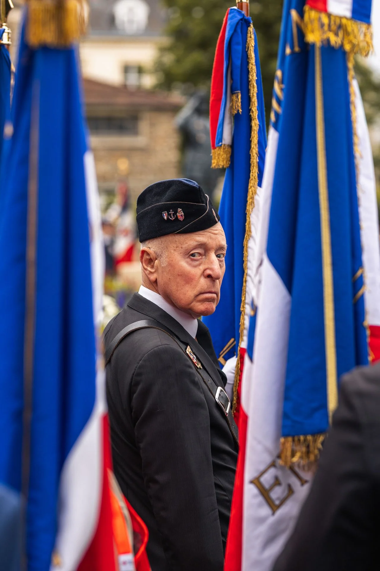 Un homme en uniforme militaire noir portant un béret noir avec des broches, debout entre des drapeaux français lors d'une cérémonie commémorative.