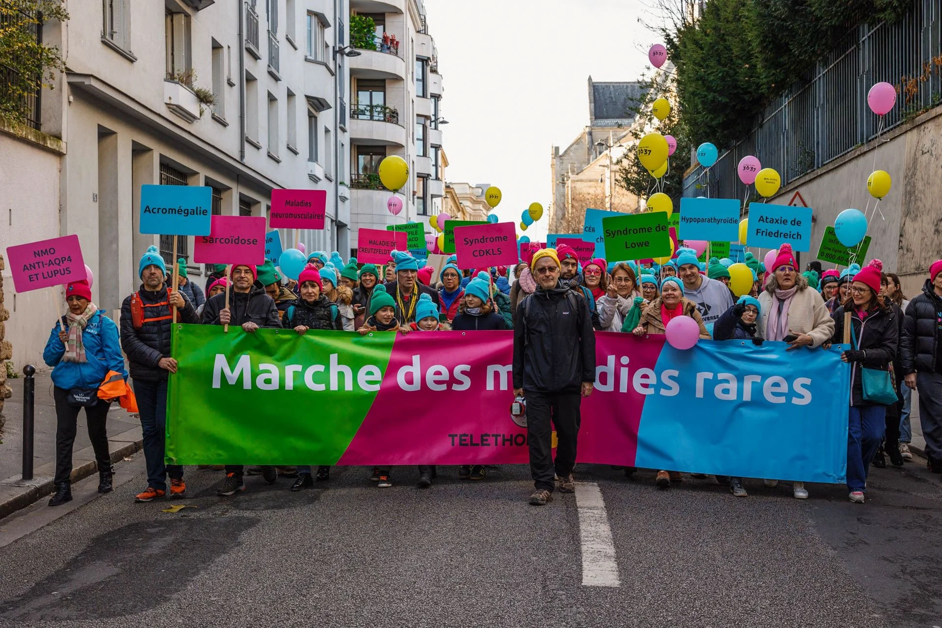 Groupe de personnes participant à une marche pour des maladies rares, portant une bannière colorée avec le texte 'Marche des maladies rares' et tenant des ballons colorés avec des noms de syndromes et de maladies