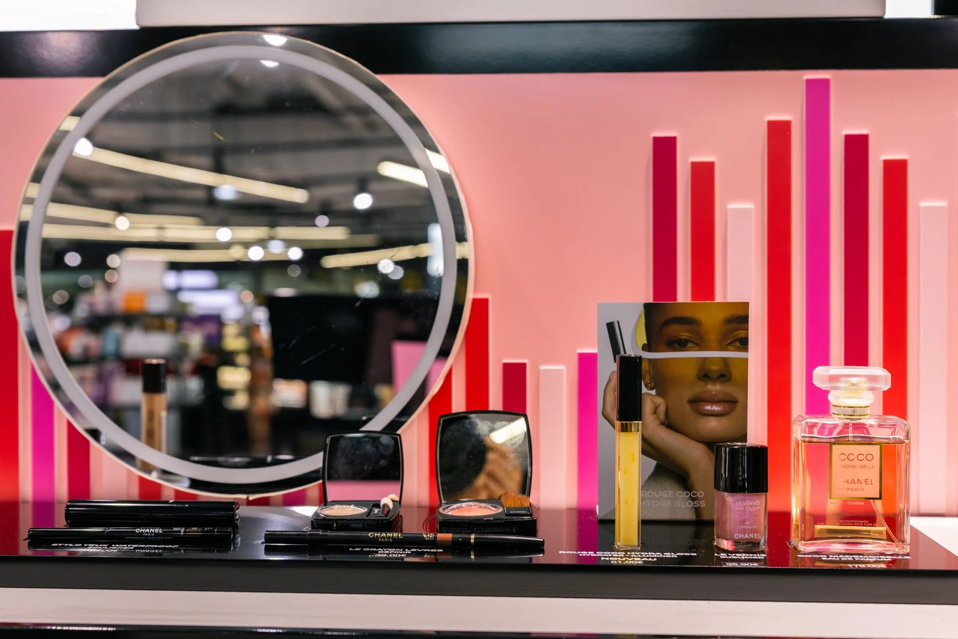 Présentation de produits cosmétiques et de maquillage dans un magasin, comprenant un miroir rond, des tubes de maquillage, un rouge à lèvres, une bouteille de parfum, et une affiche promotionnelle avec un visage de femme.