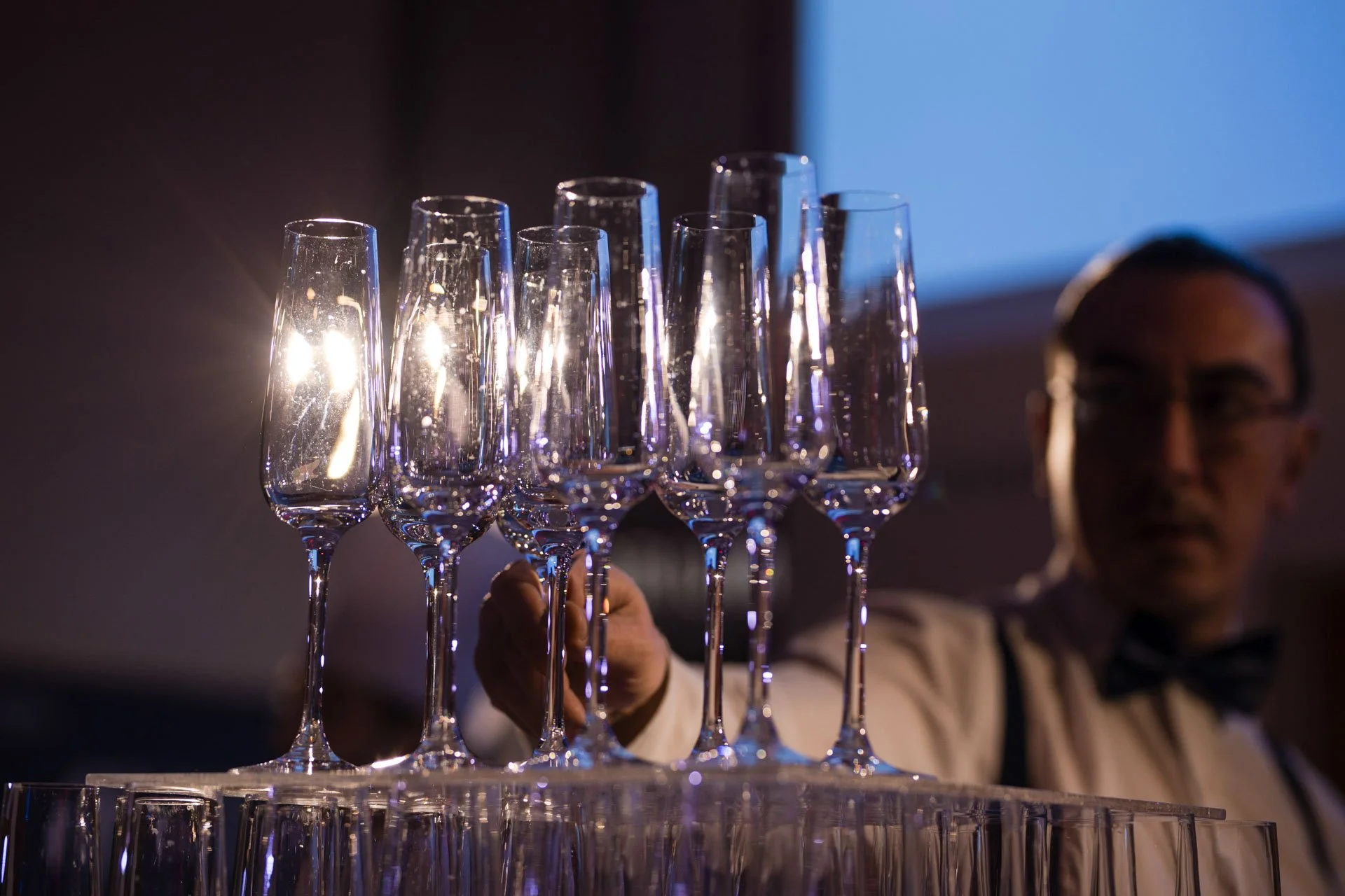 Verre à champagne éclairé de derrière, tenus par une personne, avec d'autres verres alignés en avant, dans une ambiance de fête ou de célébration, avec une personne floue en arrière-plan.