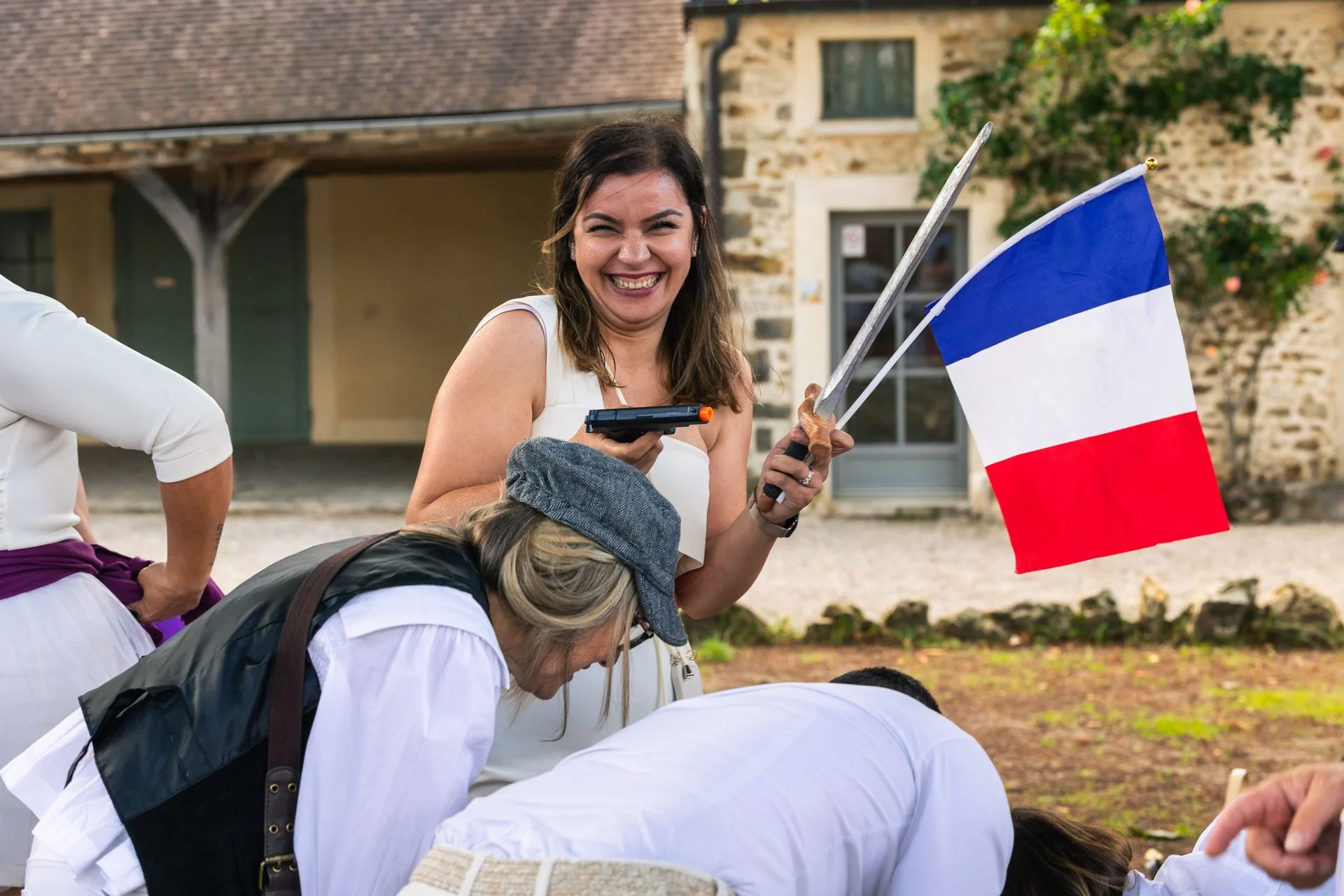 Une femme souriante tenant un drapeau français lors d'une célébration en plein air, entourée de plusieurs personnes agenouillées ou pliées en prière.