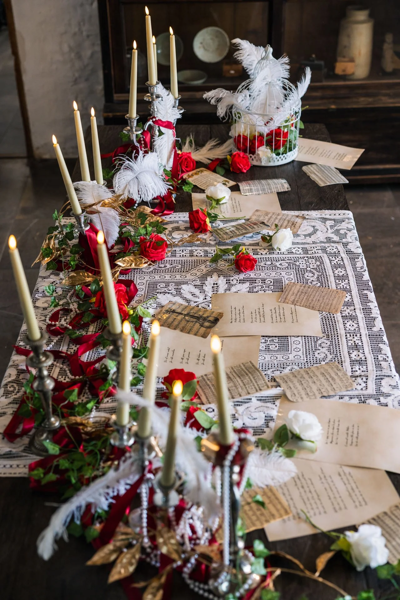 Table rustique décorée avec des chandelles allumées, des roses rouges et blanches, des plumes blanches dans une cage, papiers anciens dispersés, toile en dentelle et décorations festives.