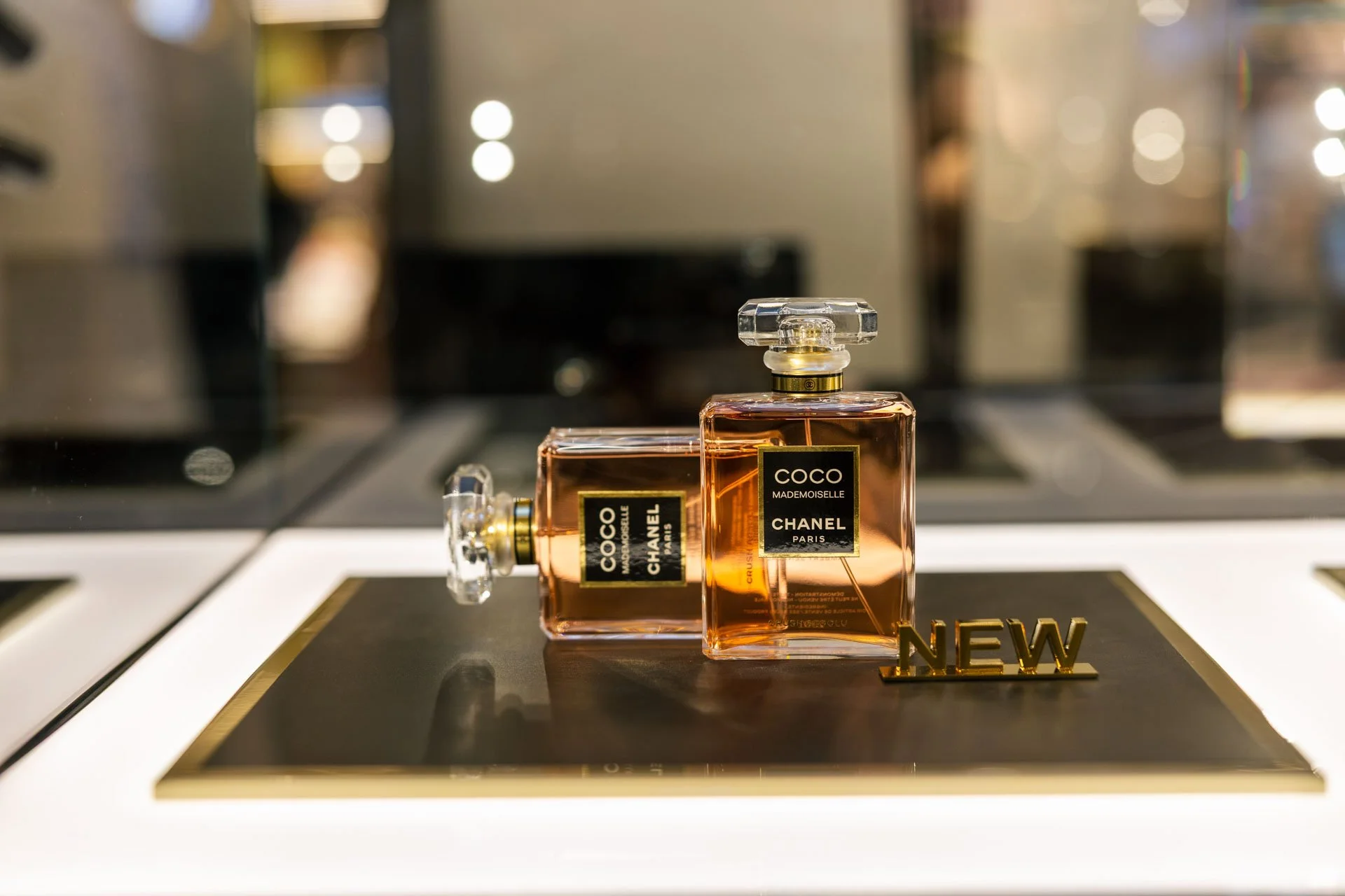 Deux flacons de parfum Chanel Coco Mademoiselle posés sur un meuble avec un décor flou en arrière-plan, et un mot 'NEW' en or au premier plan.