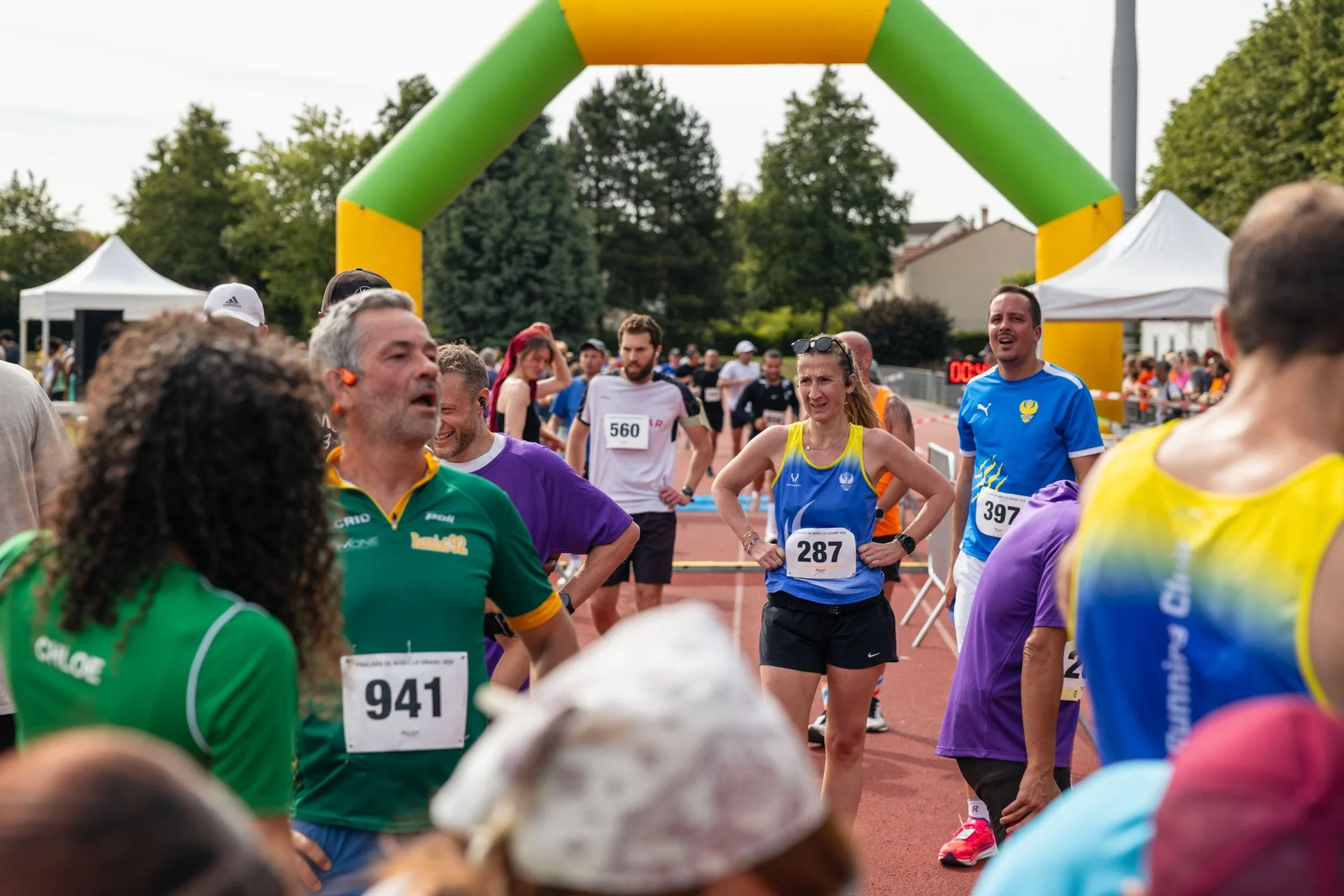 Des coureurs participent à une course, certains se serrent la main sous une arche de départ ou d'arrivée dans un stade ou une piste extérieure, avec des arbres et des tentes blanches en arrière-plan.