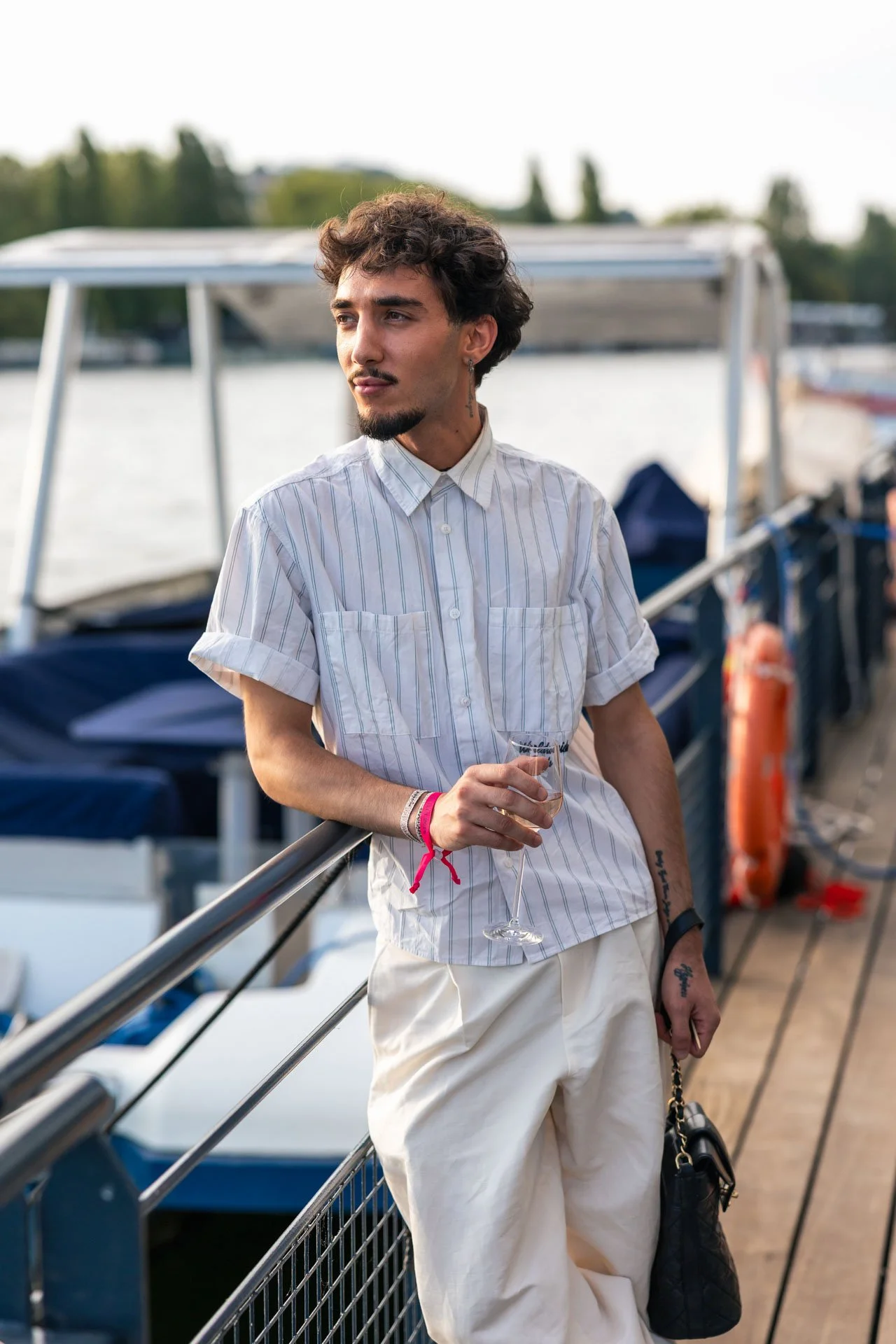 Jeune homme portant une chemise à manches courtes blanche et un pantalon ample, tenant un verre de vin et un sac à main noir, se tient près d'une barrière sur un bateau au bord de l'eau avec d'autres bateaux en arrière-plan.