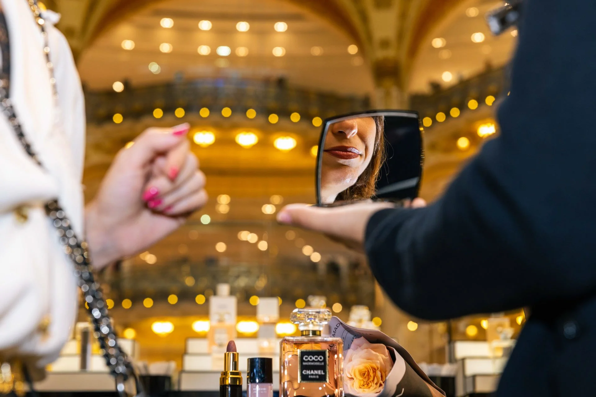 Une femme se regarde dans un miroir en faisant un essai de rouge à lèvres dans une boutique de parfums et de produits de beauté, avec des affiches et des bouteilles de parfum Chanel en avant-plan.