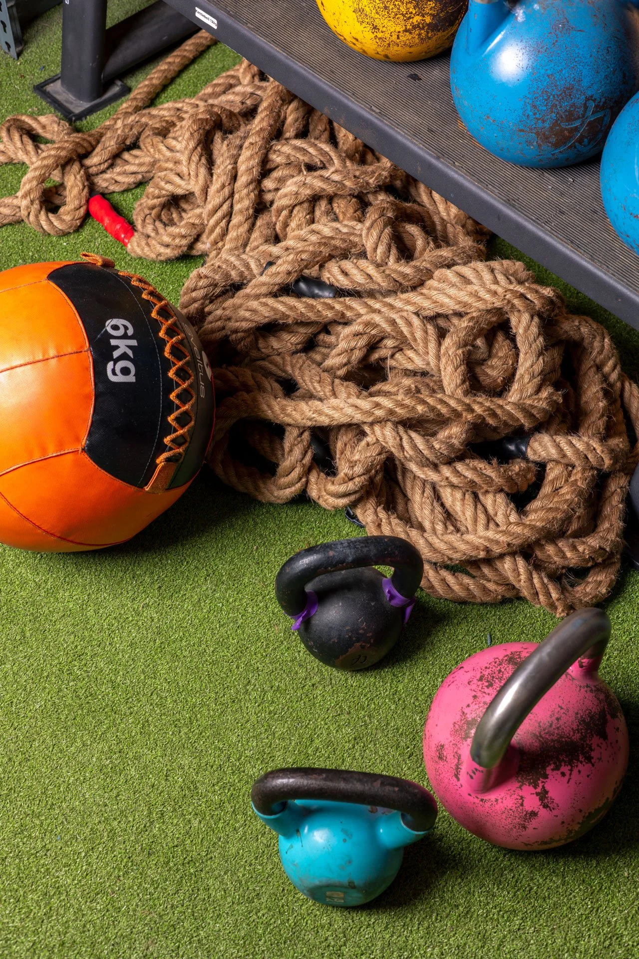Équipement de salle de sport comprenant un ballon de football orange et noir, des chaînes épaisses, des kettlebells colorés (rose, bleu, noir), et des kettlebells bleus et jaunes sur une étagère.