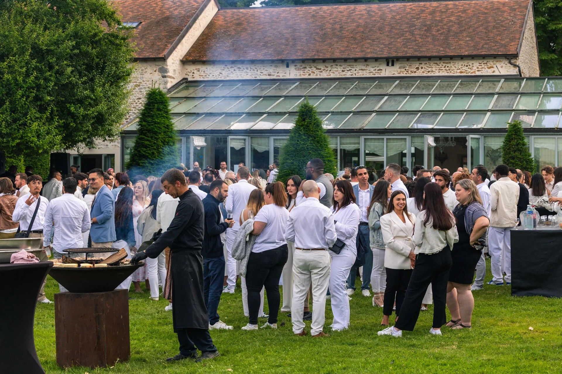 Gathering en plein air devant une villa avec un espace fermé en verre, beaucoup de personnes habillées en blanc, certains cuisinant sur un barbecue.