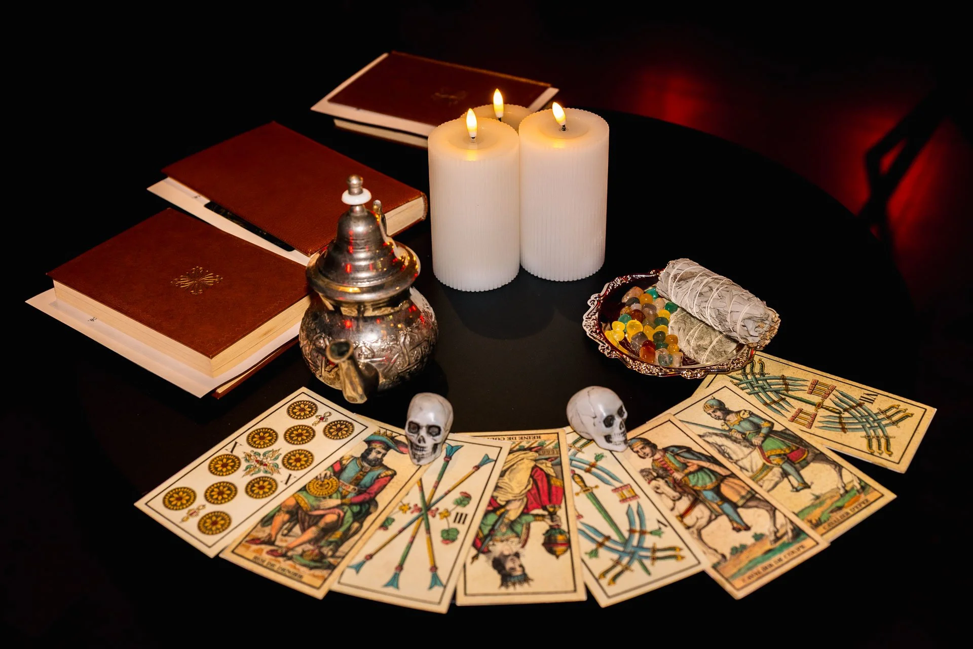 Jeu de tarot avec cartes étalées, petits crânes en plastique, bougies allumées, livres, pièces de monnaie, un flacon en métal, un paquet de sauge, et une assiette de pierres de décoration sur une table noire.
