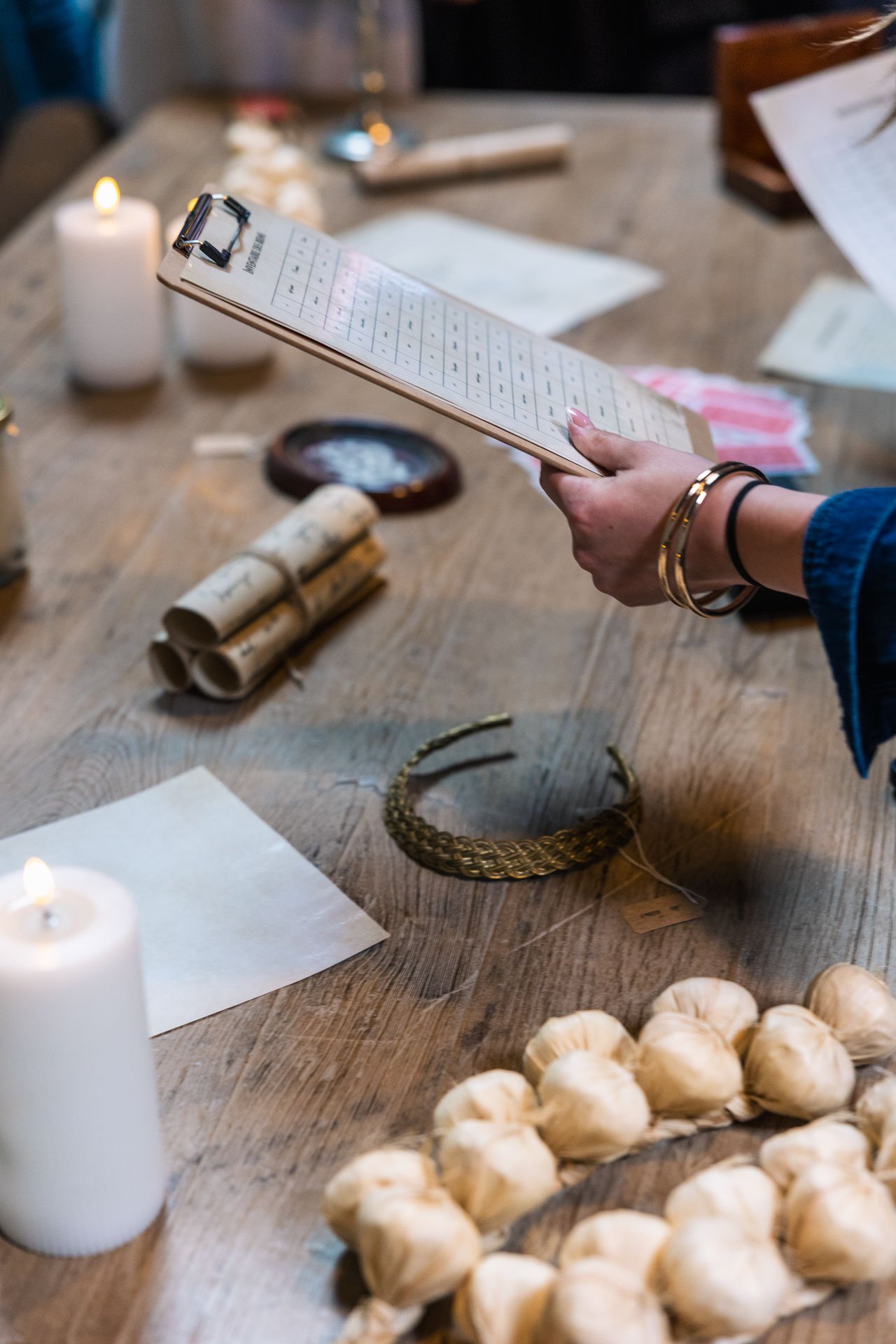 Personne tenant une fiche de calendrier à la main sur une table en bois, entourée de bougies, papiers, rouleaux de paperasserie, une couronne en métal, des petits sacs de pommes de terre, et une vaisselle vintage