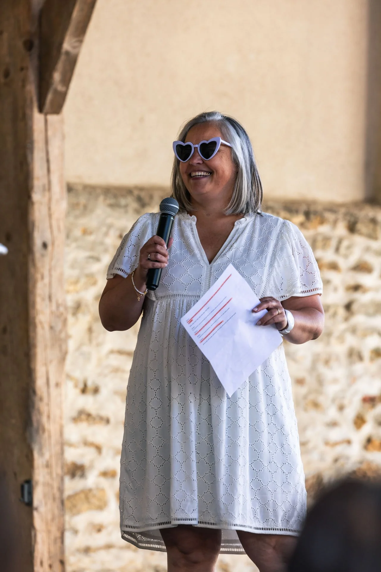 Une femme souriante portant des lunettes en forme de cœurs et une robe blanche, tenant un micro et une feuille de papier, se tenant devant un mur en pierre.