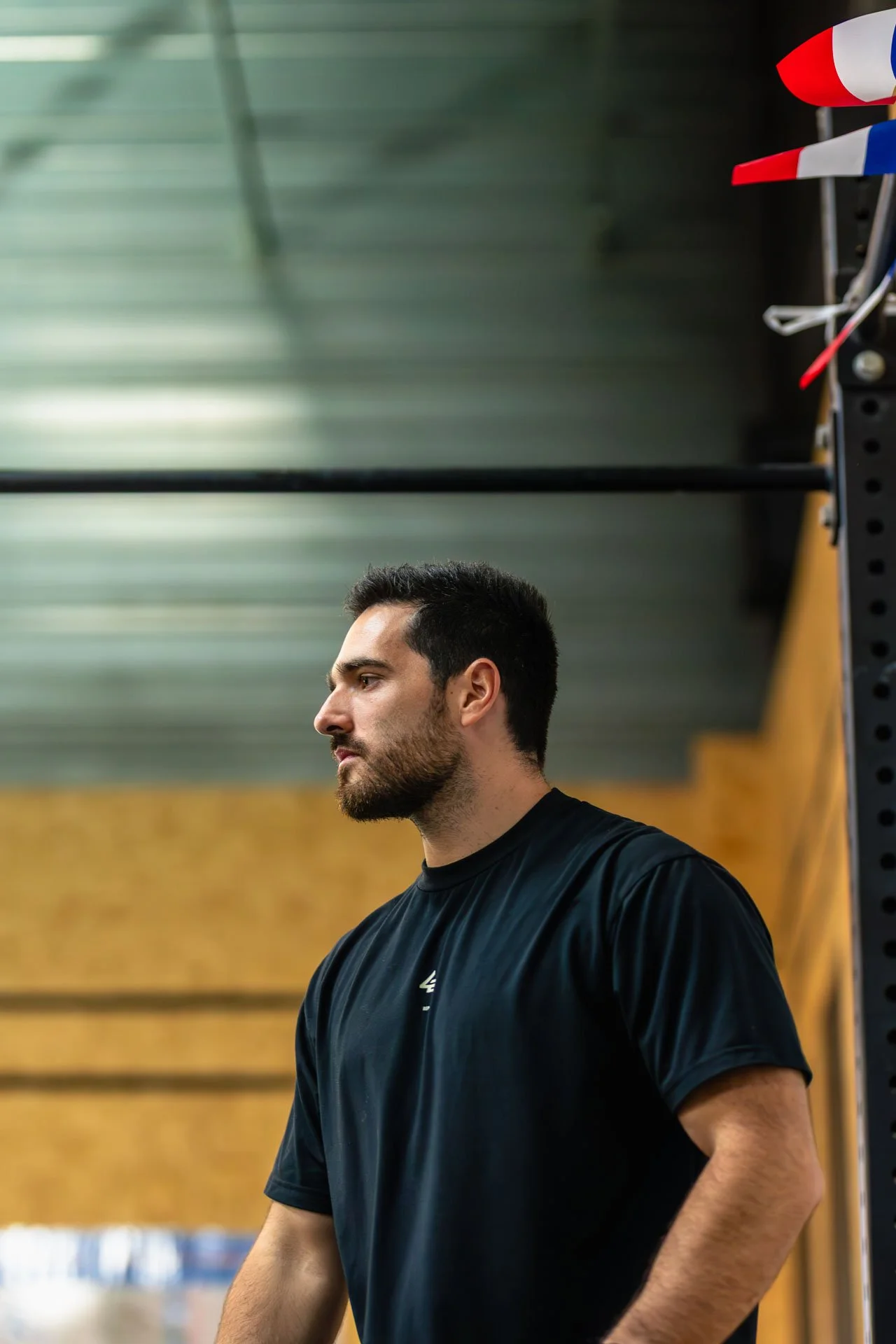 Un homme musclé, portant un t-shirt noir, regarde pensivement dans une salle de sport avec des murs en bois, un plafond en métal et des bandes de résistance suspendues