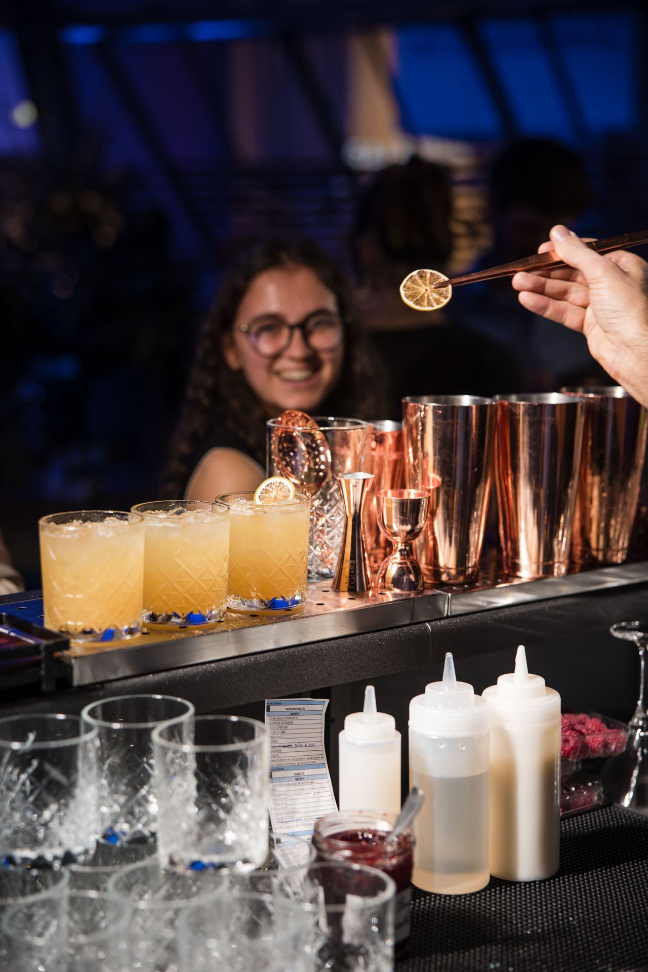 Un bar avec des cocktails colorés, des shakers en cuivre, des bouteilles de sirops et des verres en cristal. Une femme souriante en arrière-plan
