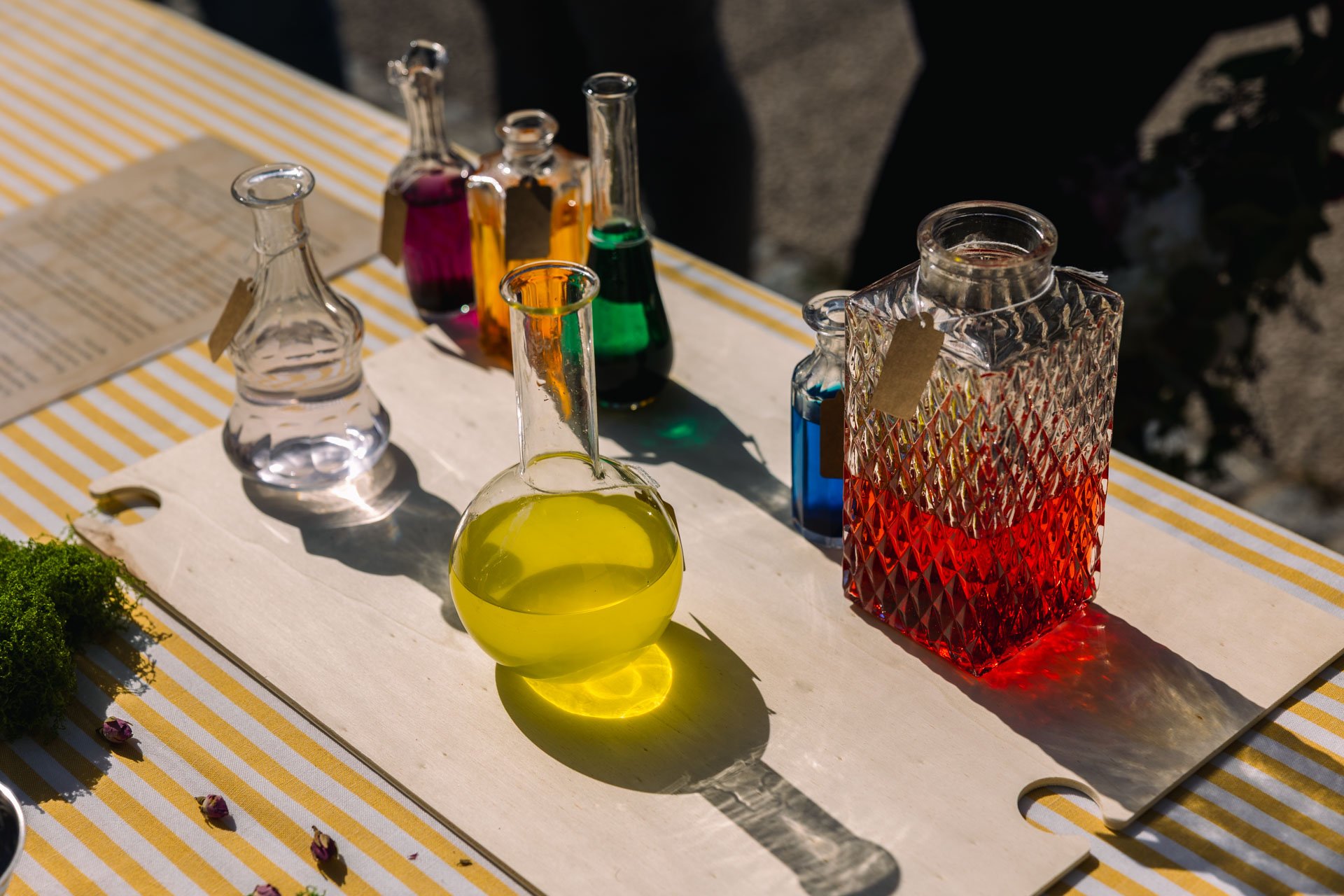 Verres en forme de bouteille contenant des liquides colorés, exposés sur une planche en bois sur une table avec un tissu à rayures jaunes et blanches.