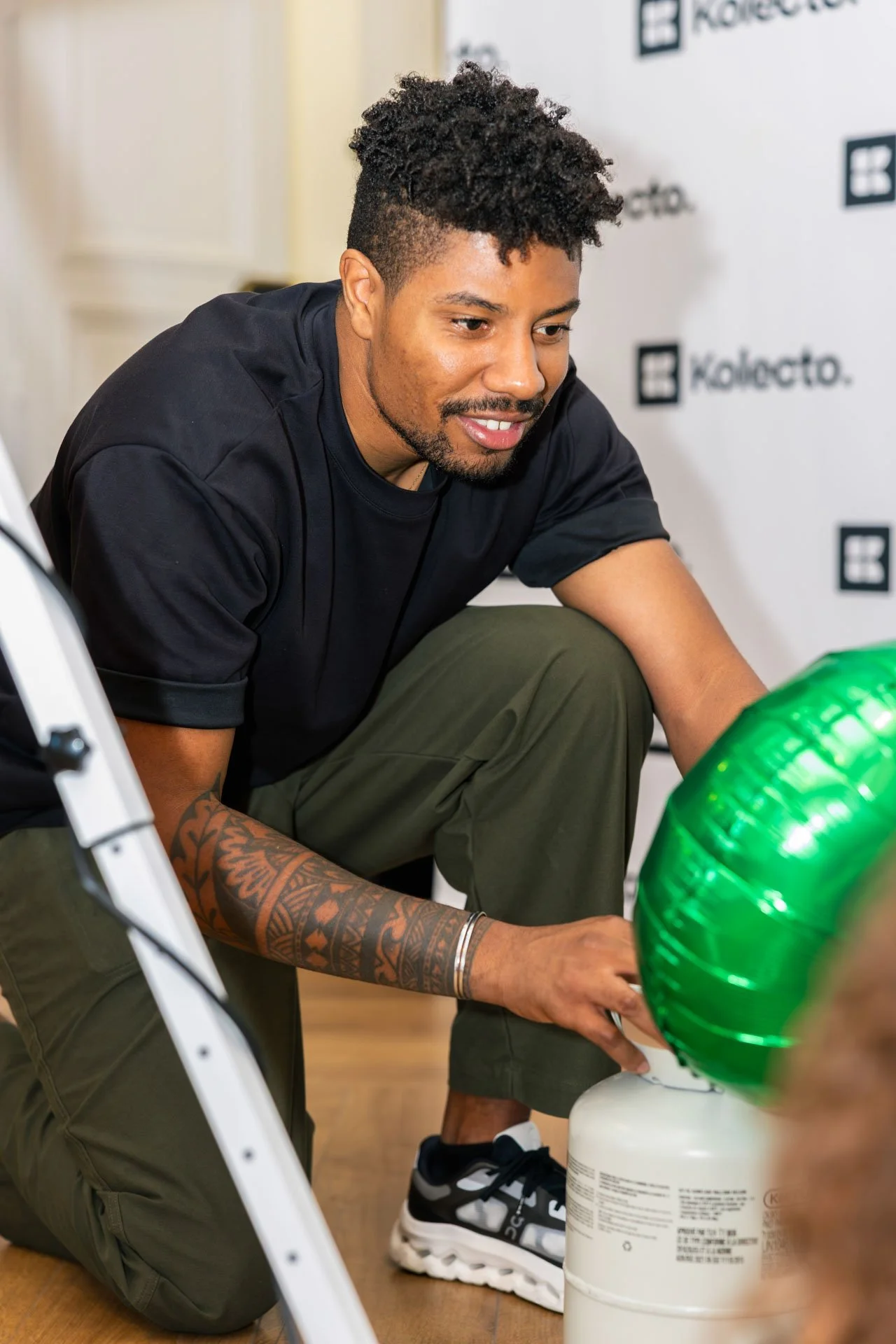 Un homme assis, en train de faire de la sculpture avec un ballon vert, avec un fond blanc avec des logos.