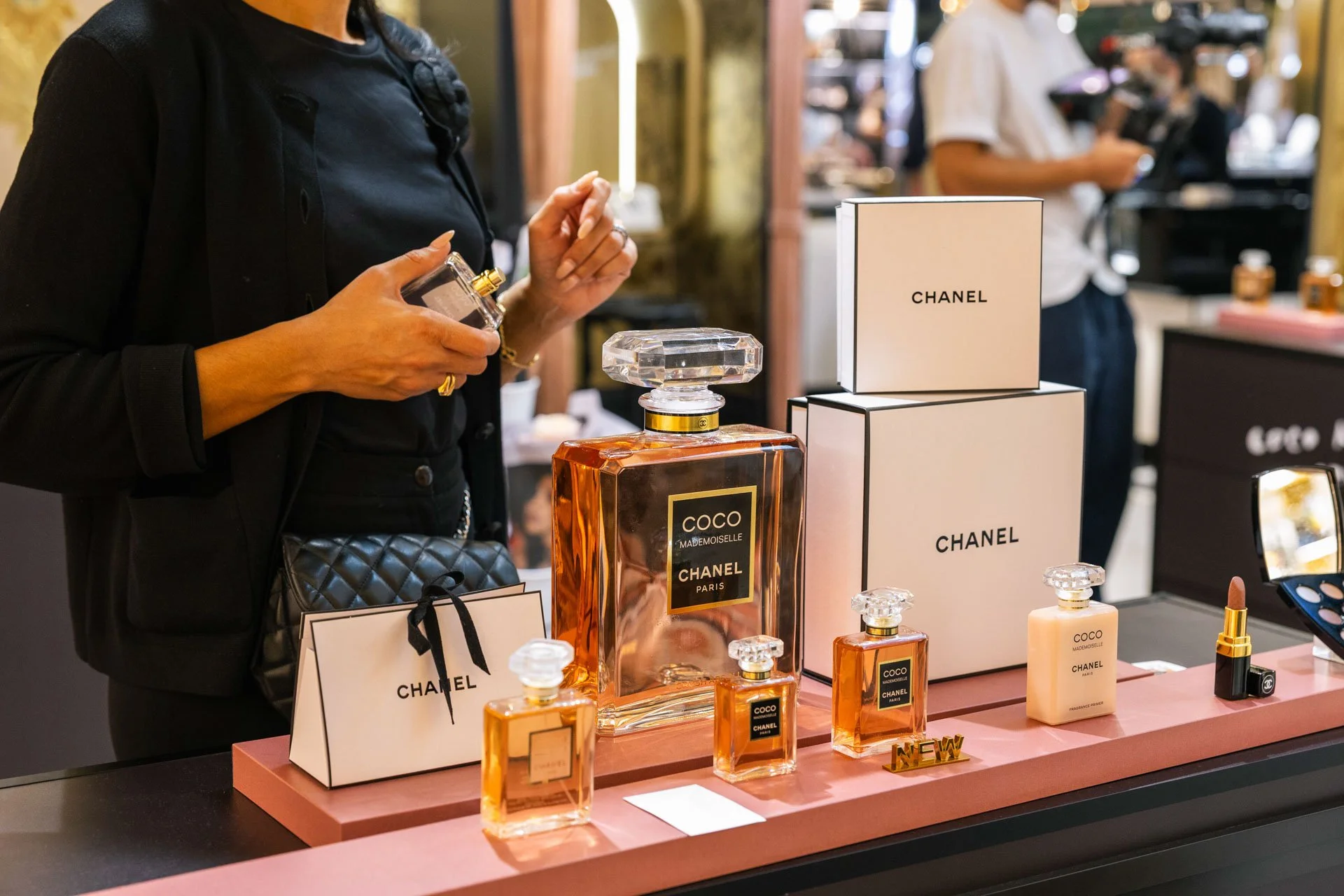 Une personne achète des parfums Chanel. Sur la table, plusieurs flacons de parfum, boîtes et accessoires de la marque Chanel. En arrière-plan, d'autres personnes dans un magasin.