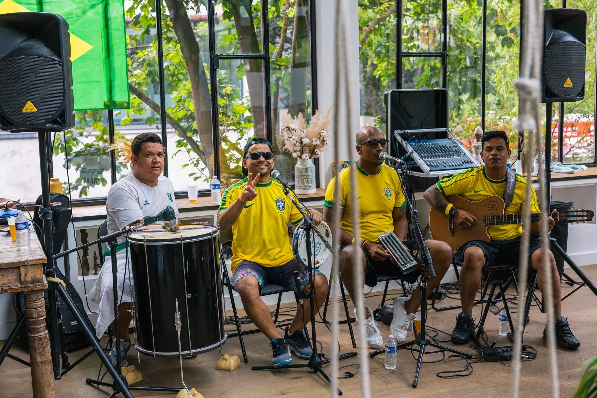 Groupe de musiciens jouant dans un espace intérieur lumineux. Ils portent des maillots de football brésiliens, certains jouent du tambour, guitare et d'autres instruments, avec équipement de sonorisation autour d'eux.